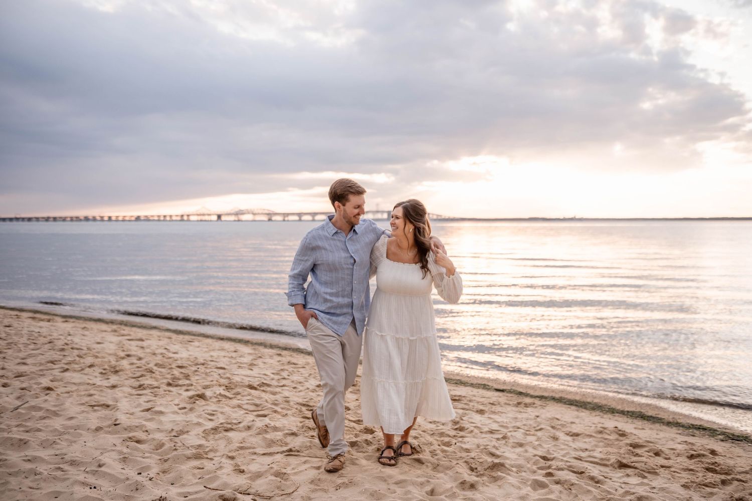 Couple walking hand in hand on sandy beach at sunset with bridge visible in distance.