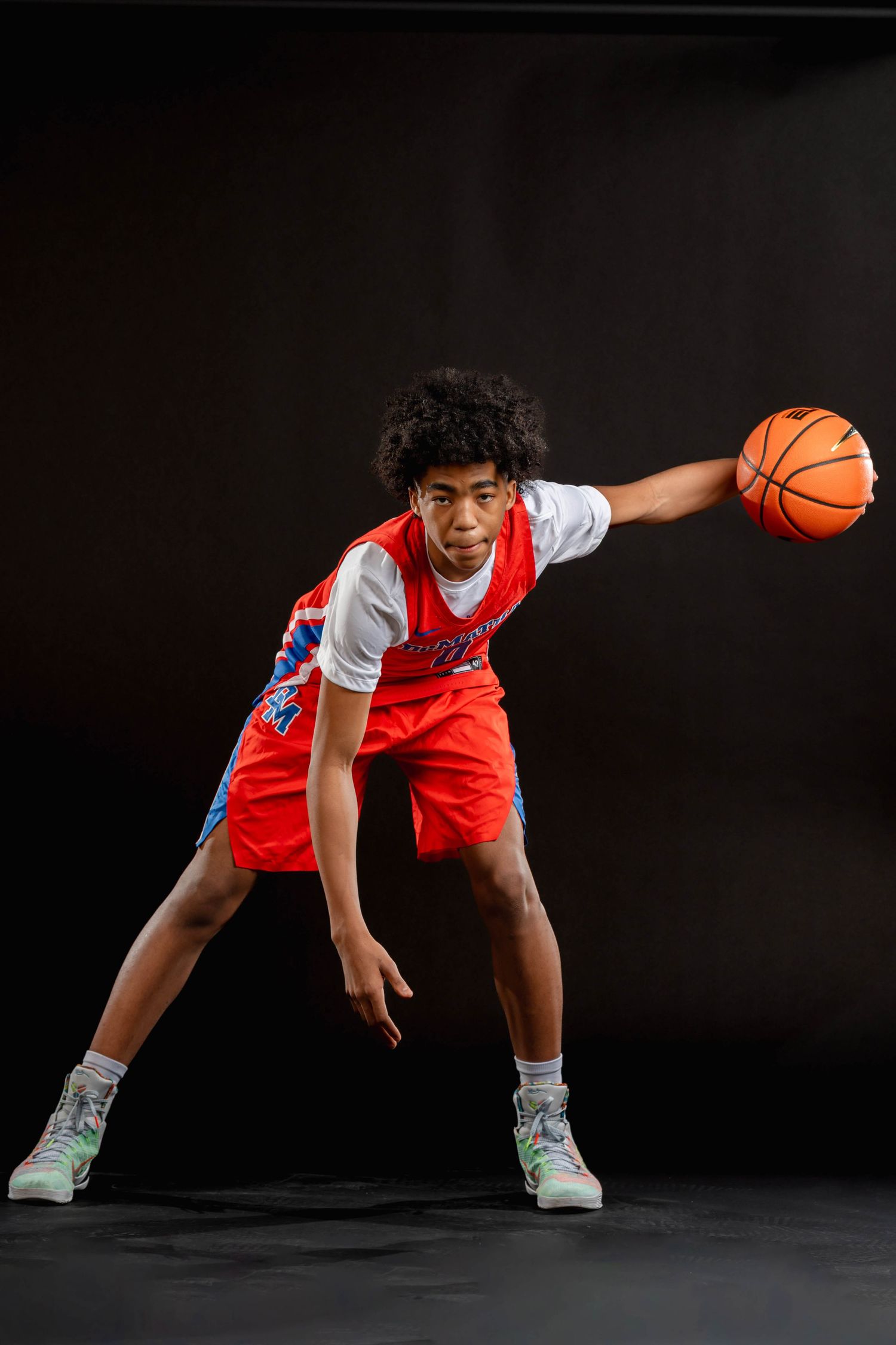 Young basketball player in red DeMatha uniform dribbles ball in dark studio.