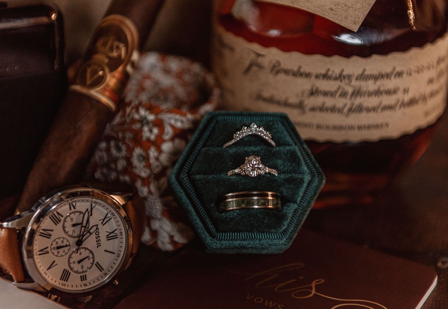 Wedding flat lay with diamond engagement ring and band in velvet box, beside a watch, cigar, and bourbon bottle.