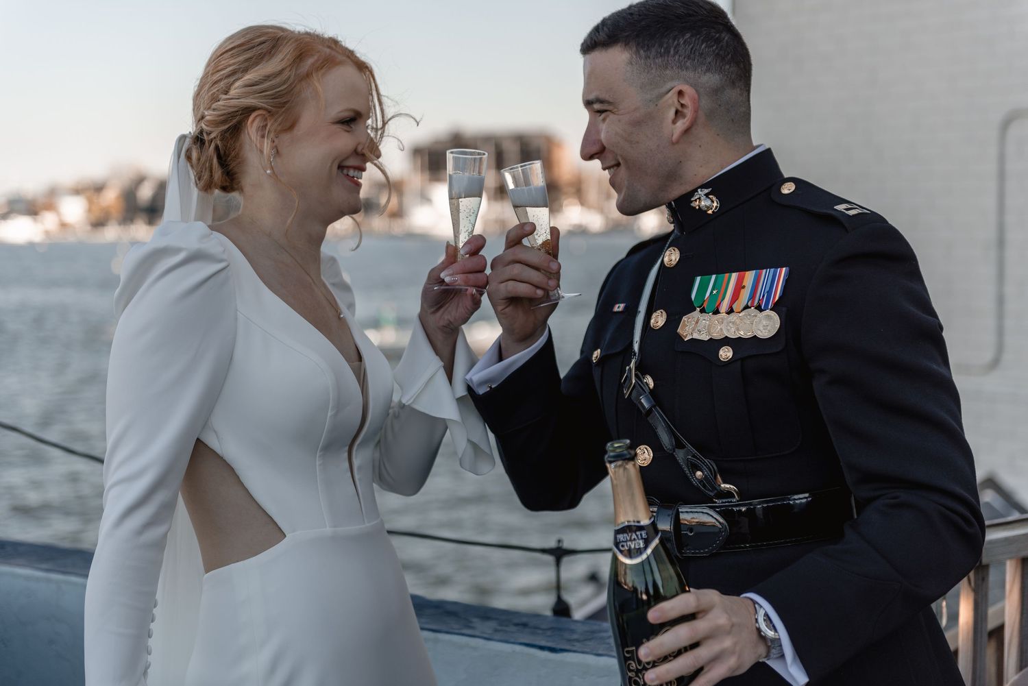 Newlyweds toast with champagne glasses aboard a boat, groom in military uniform at waterfront wedding.
