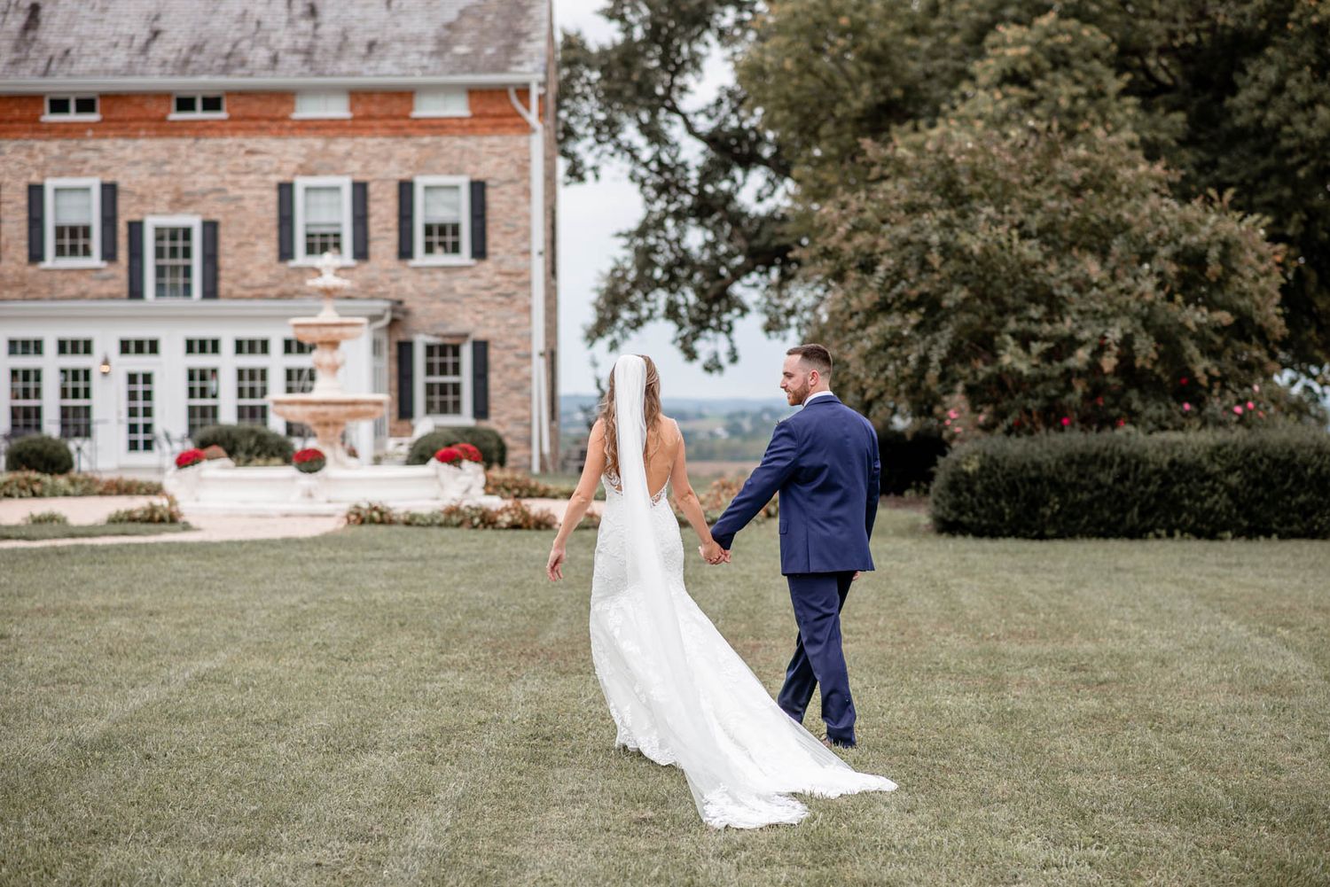 A bride and groom walk hand in hand across a lush lawn toward an elegant brick estate on their wedding day.