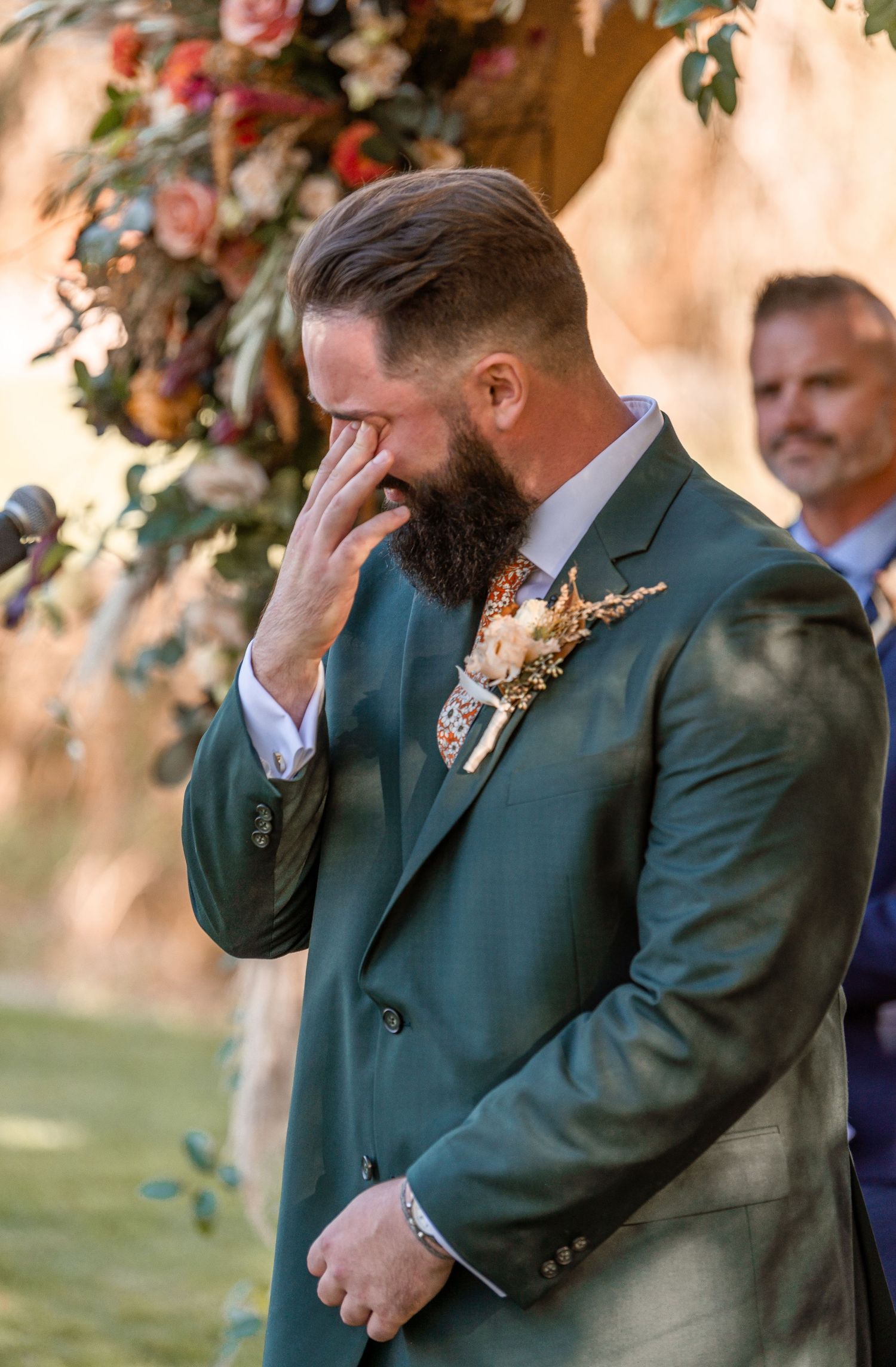 Emotional bearded groom in a green suit wipes tears at the altar beside a colorful floral wedding arch.