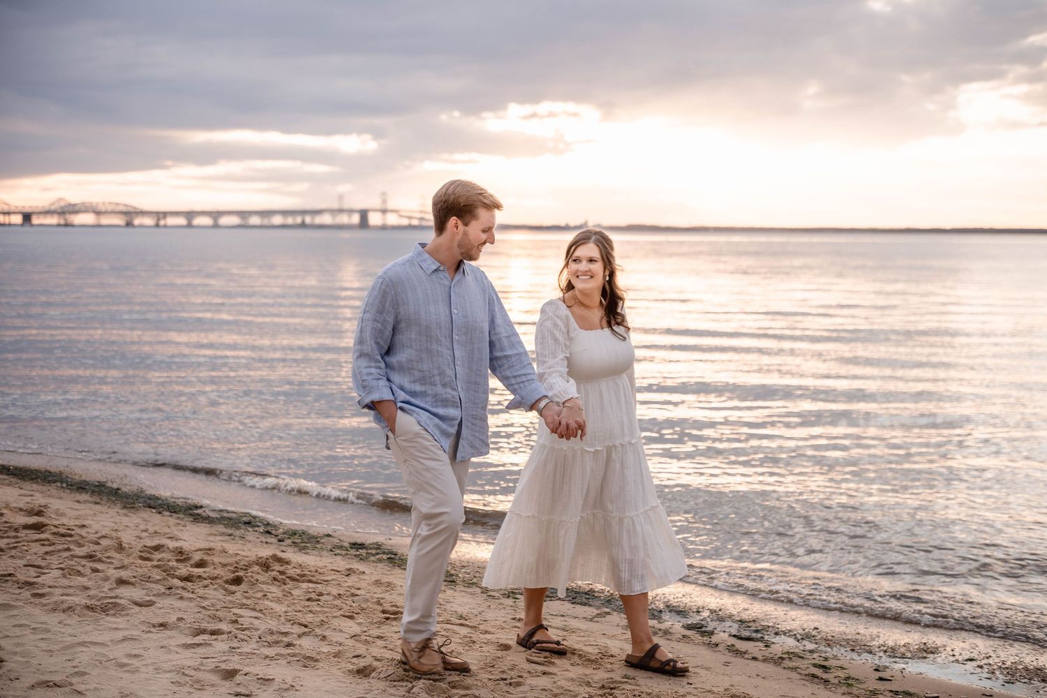 Couple walks hand in hand on sandy beach at sunset with bridge and colorful sky in background.