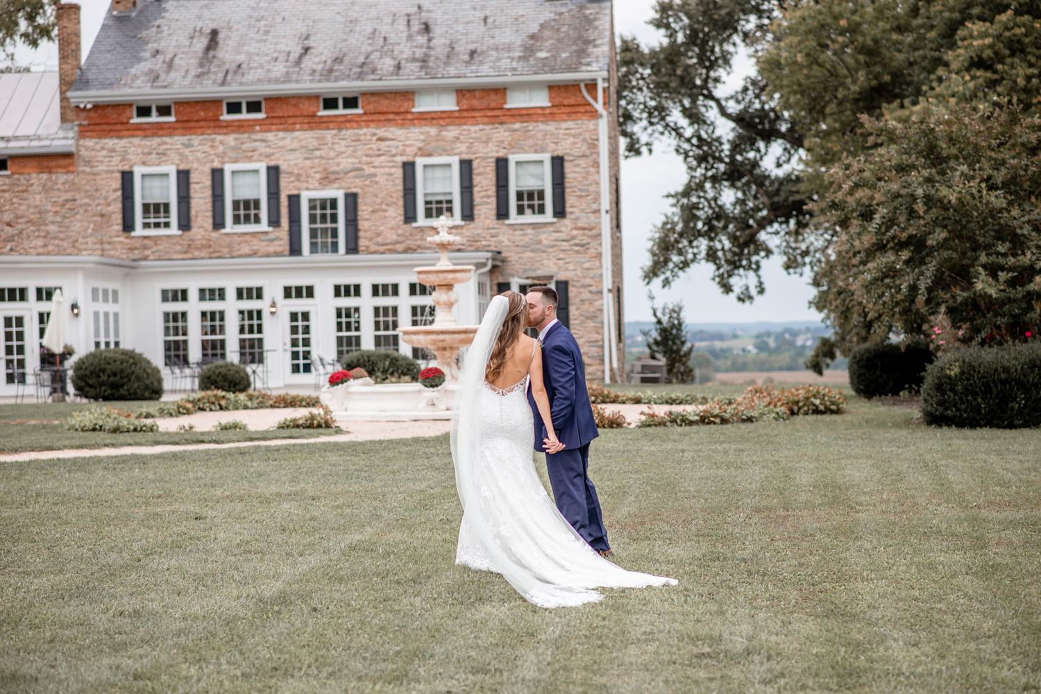 Bride and groom share a romantic moment on a manicured lawn in front of a grand historic brick manor.