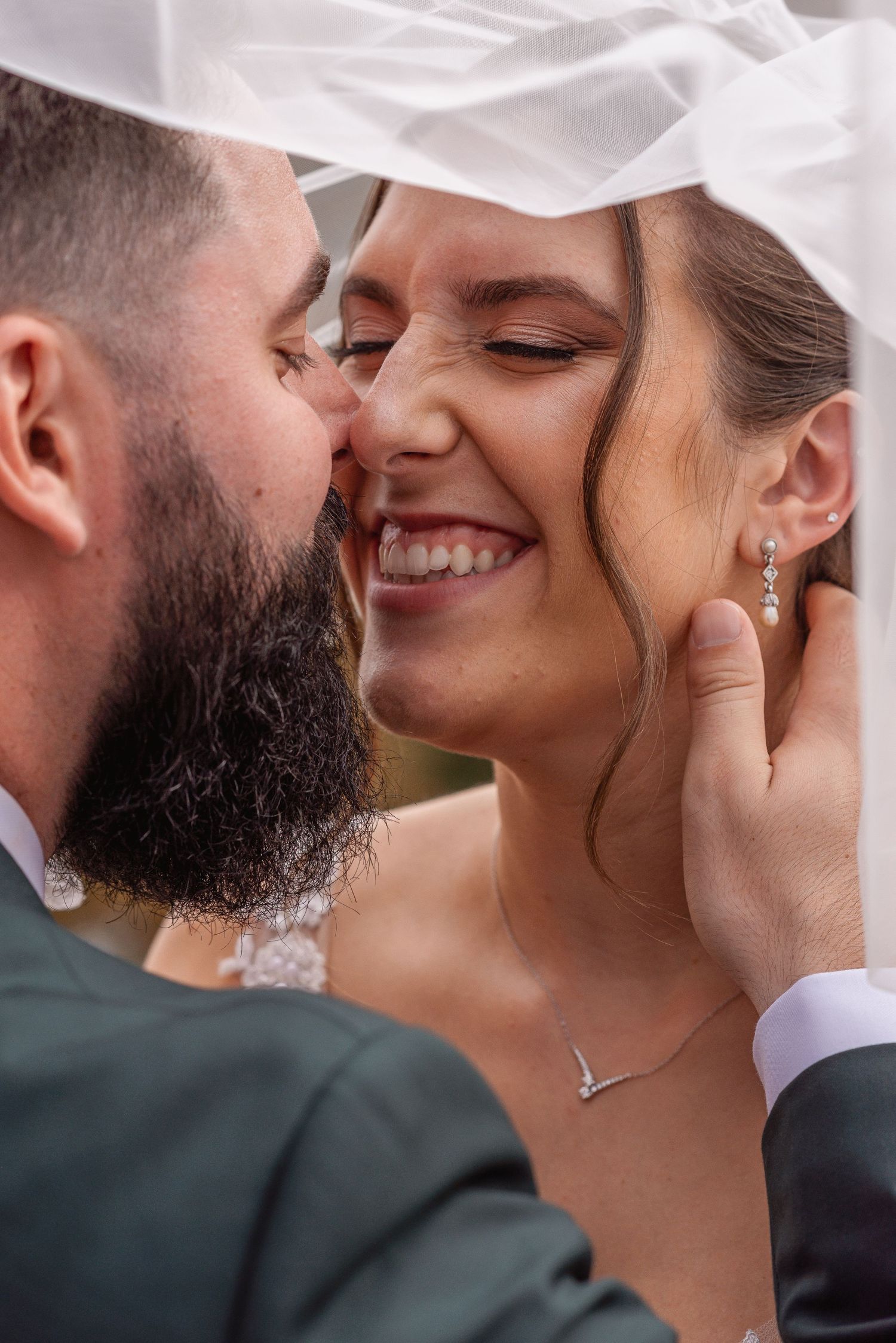 Groom tenderly cupping bride's face under a veil as she smiles with eyes closed at their wedding.