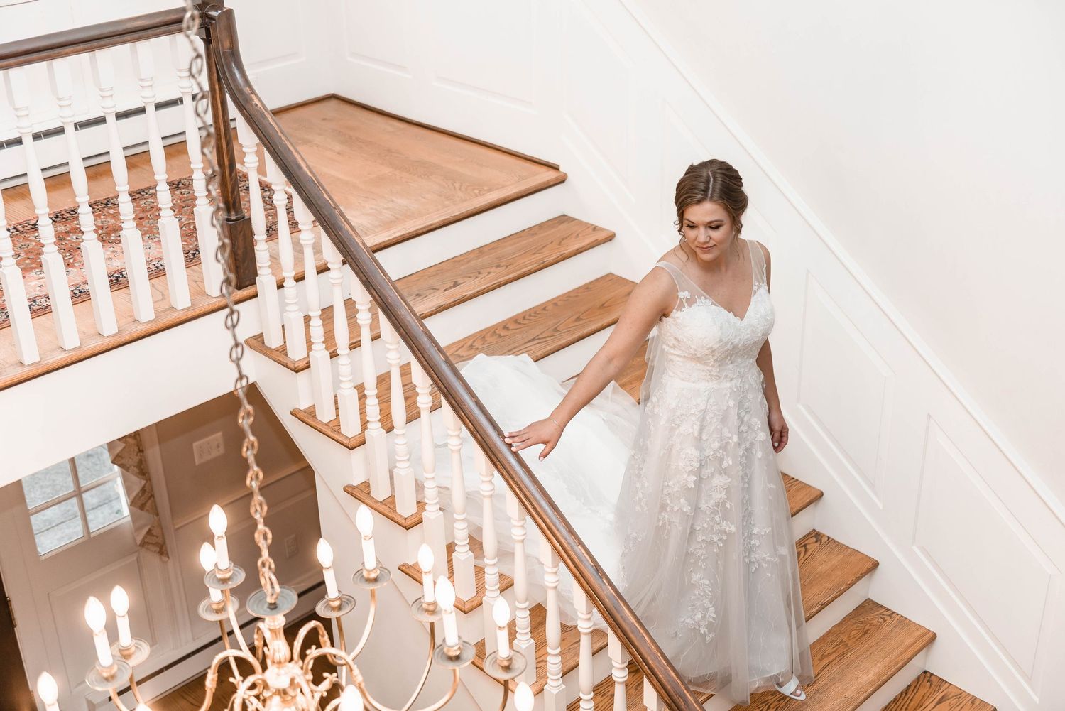 Elegant bride in lace wedding gown descending grand staircase with chandelier below.
