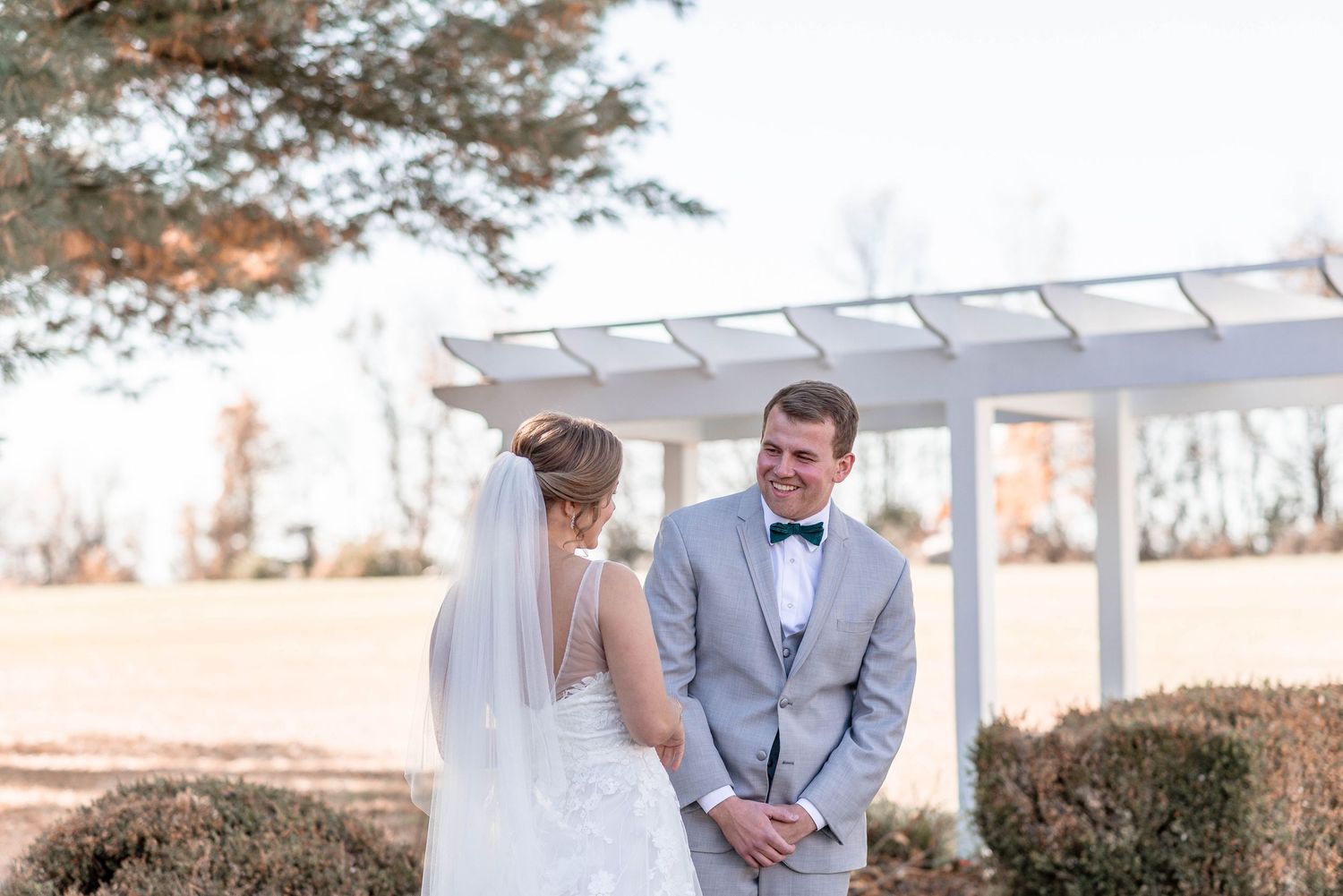 Groom in gray suit smiles at bride during emotional first look near a white garden pergola.