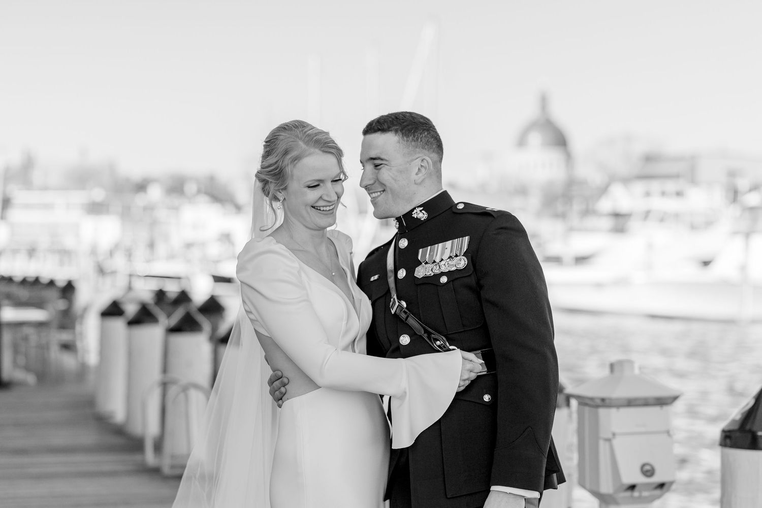Black and white photo of smiling bride and Marine groom embracing at waterfront marina on wedding day.