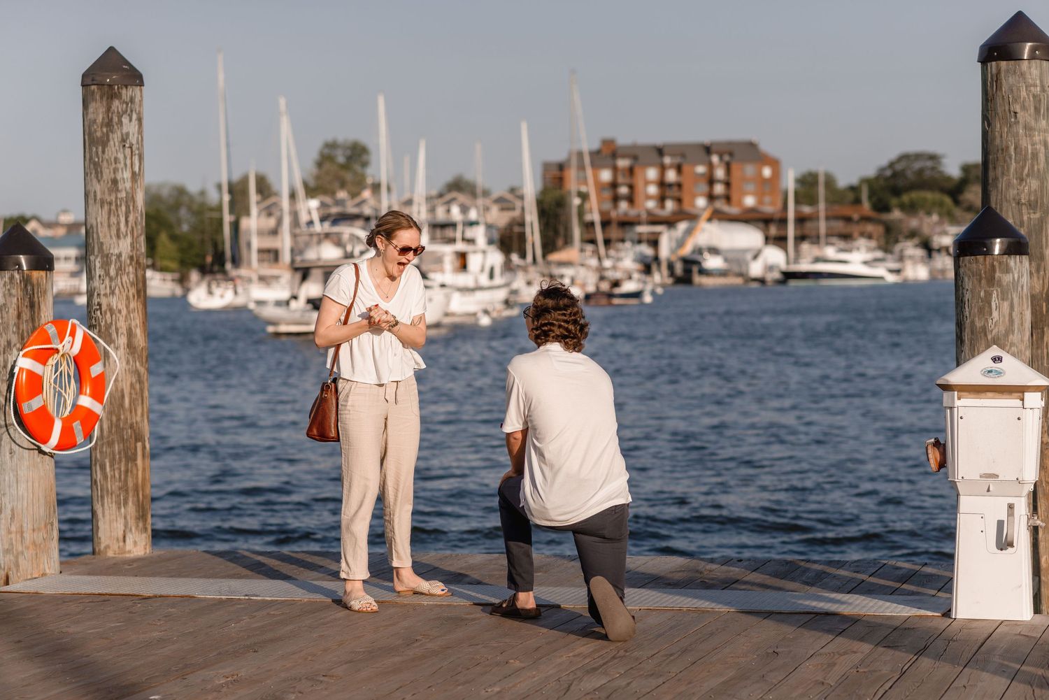 Man kneeling on a waterfront dock proposing to his girlfriend with sailboats in the background.