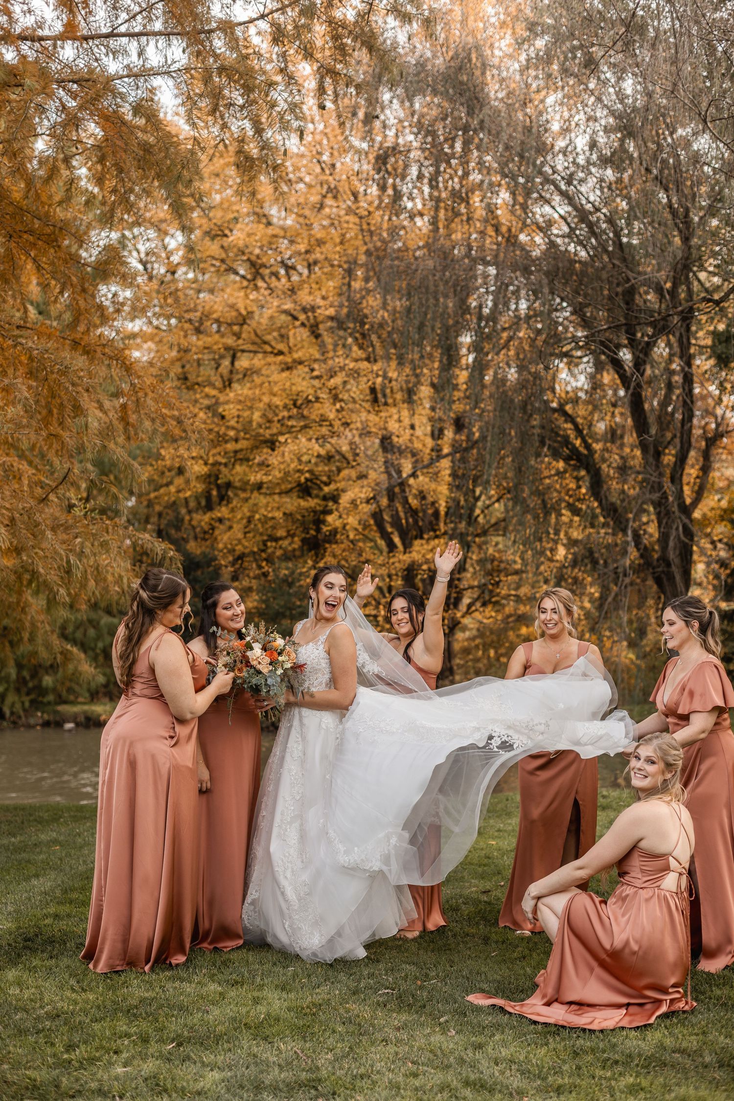 Bridesmaids in terracotta dresses joyfully lifting bride's gown outdoors with golden autumn trees behind.