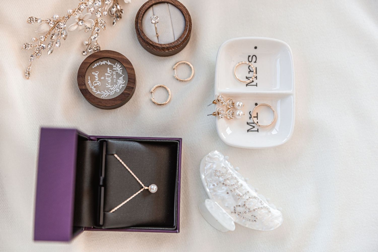 Elegant wedding flat lay featuring watch, rings, Mr and Mrs dishes, and pearl earrings on white fabric.