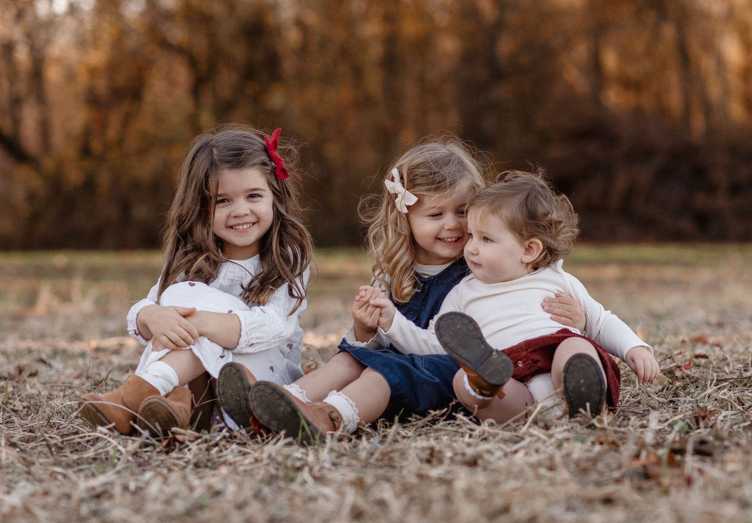 Three young children sitting together on autumn grass, smiling and laughing in coordinated white outfits.