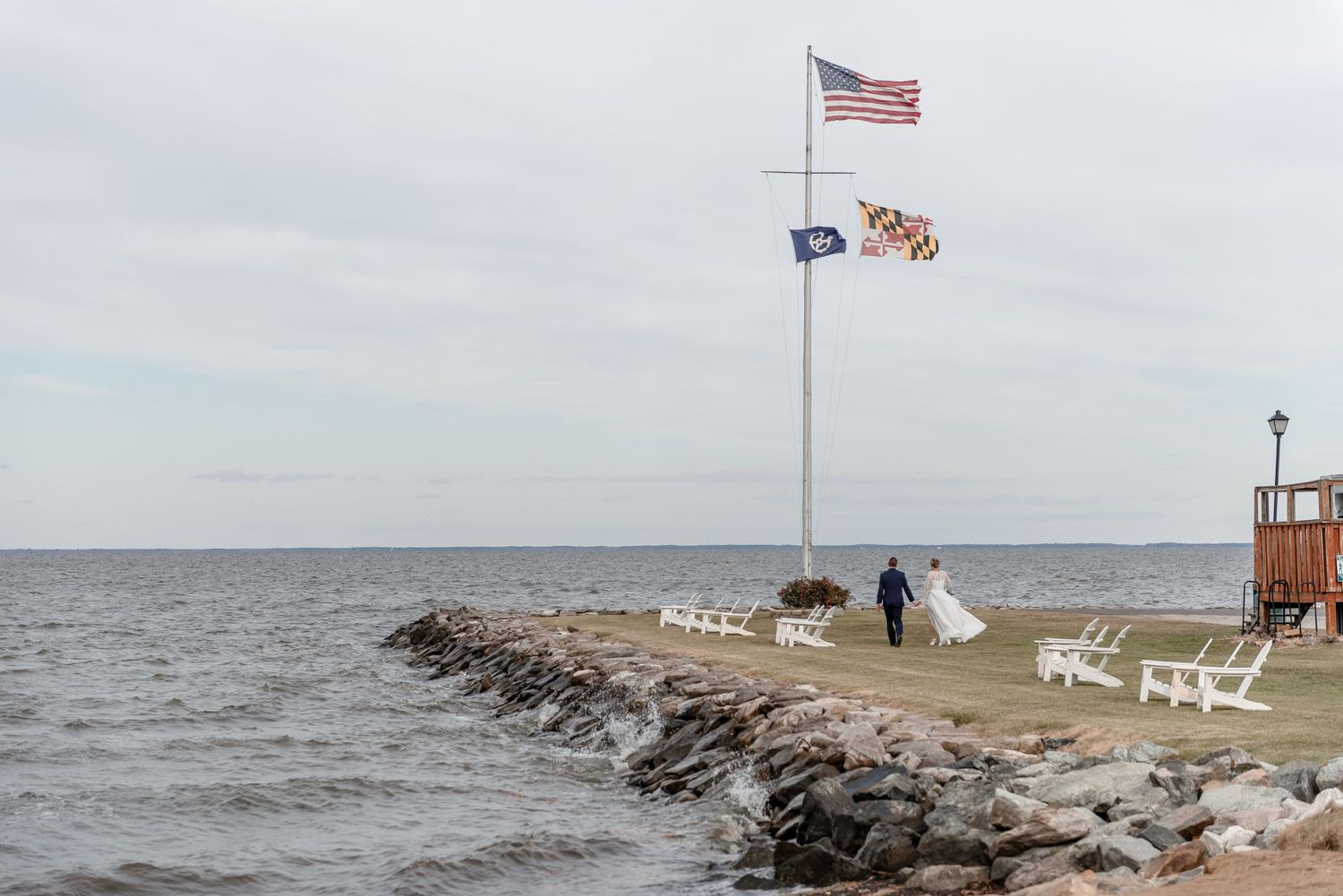 Bride and groom walk near flagpole with American and Maryland flags by waterfront.