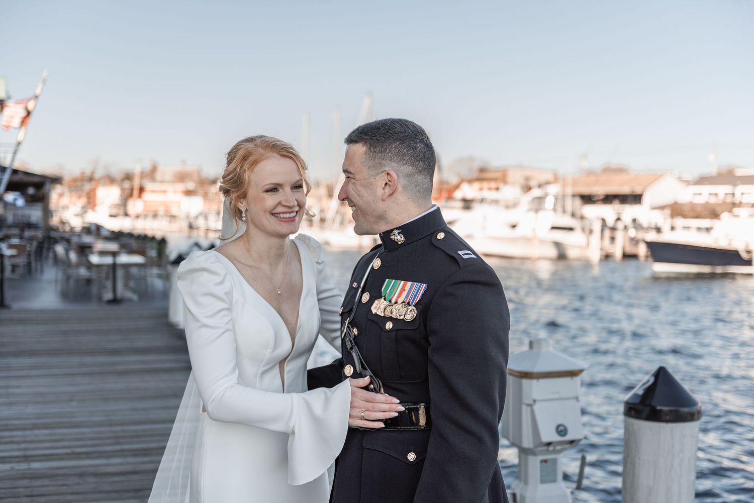 Bride and groom in military dress uniform share a romantic moment on a sunny marina dock.