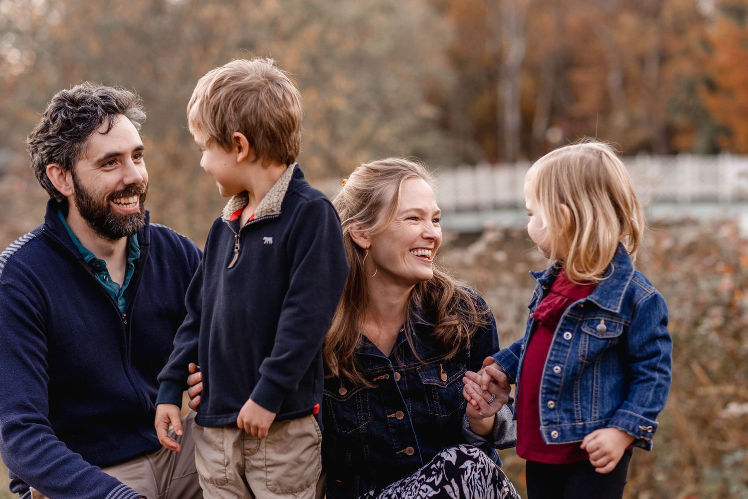 A happy family of four laughs together outdoors surrounded by colorful autumn foliage in a park.