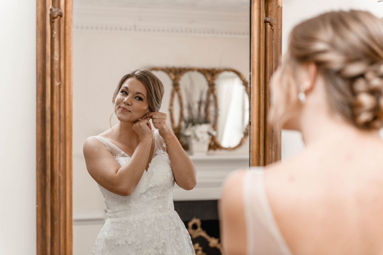 Bride in lace wedding gown adjusts earring while gazing into an ornate wooden framed mirror.