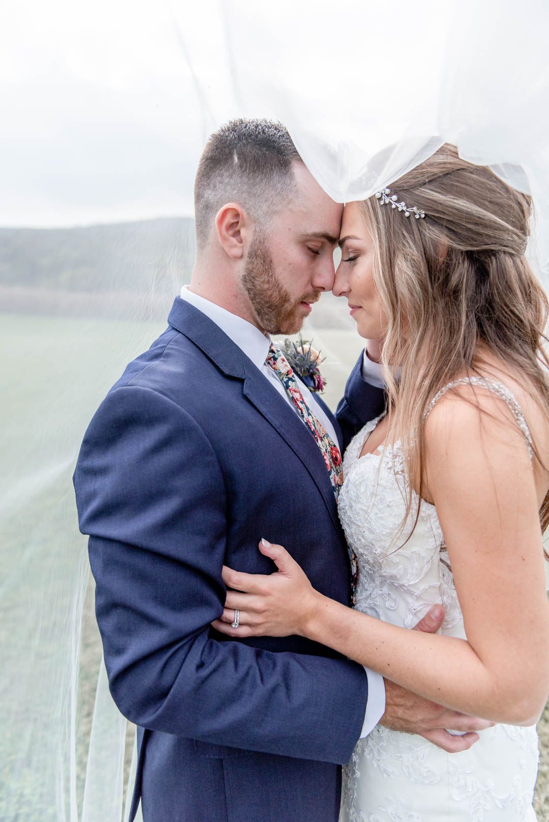 Bride and groom share an intimate moment under a veil with foreheads touching at outdoor wedding.