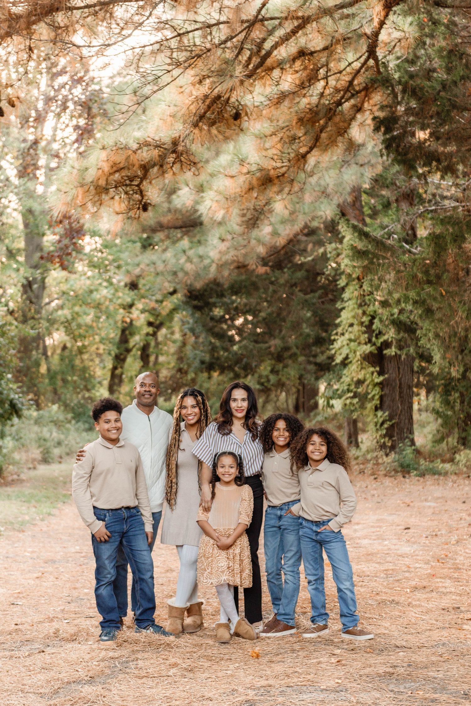 A happy family of seven posing together on a leaf-covered path beneath colorful autumn trees in a park.