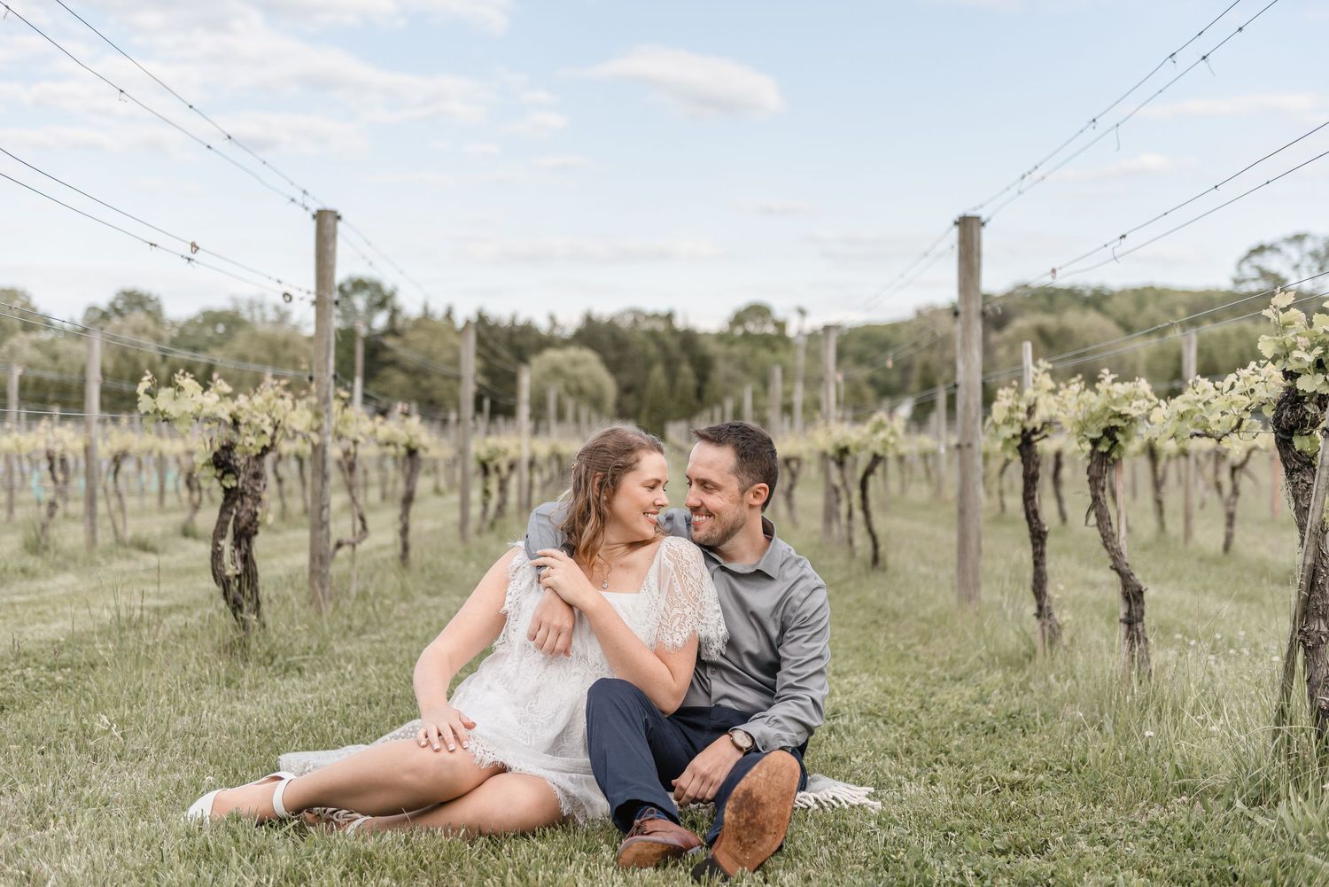 Couple sitting together in a vineyard, smiling and embracing on a grassy path between vine rows.