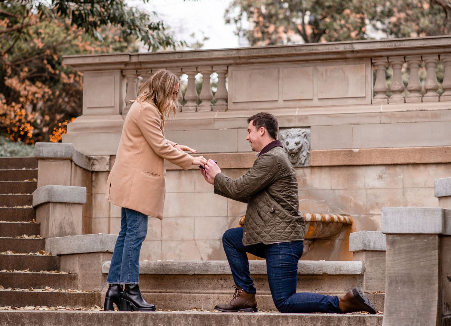 Man proposing on one knee on stone steps while holding womans hands at an outdoor landmark.