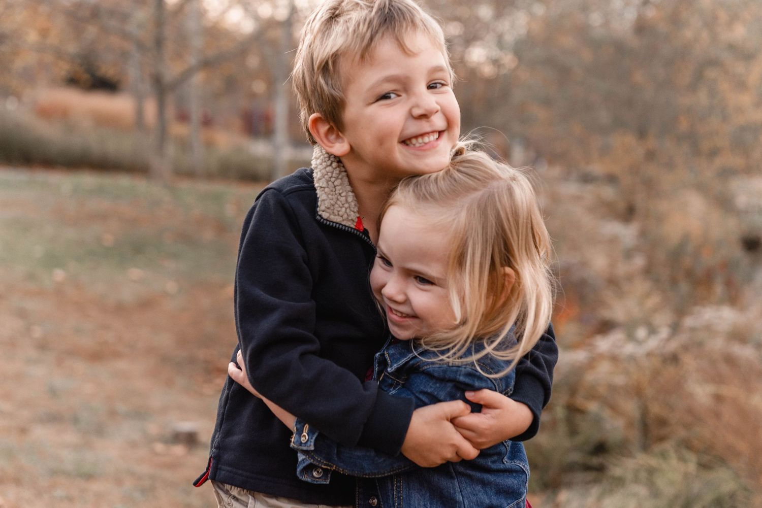 Young boy hugging little girl in denim jacket, both smiling during outdoor fall sibling photo session.