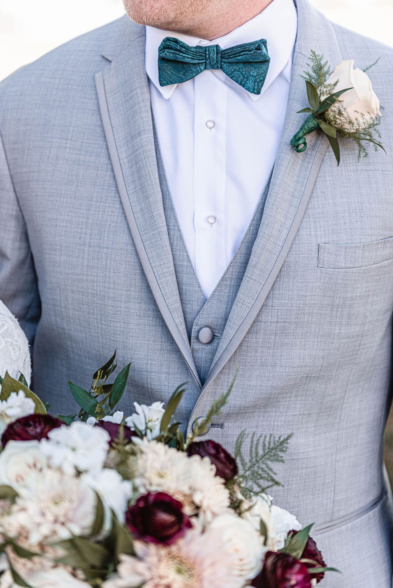 Groom in gray suit with teal bow tie and boutonniere holding bridal bouquet.