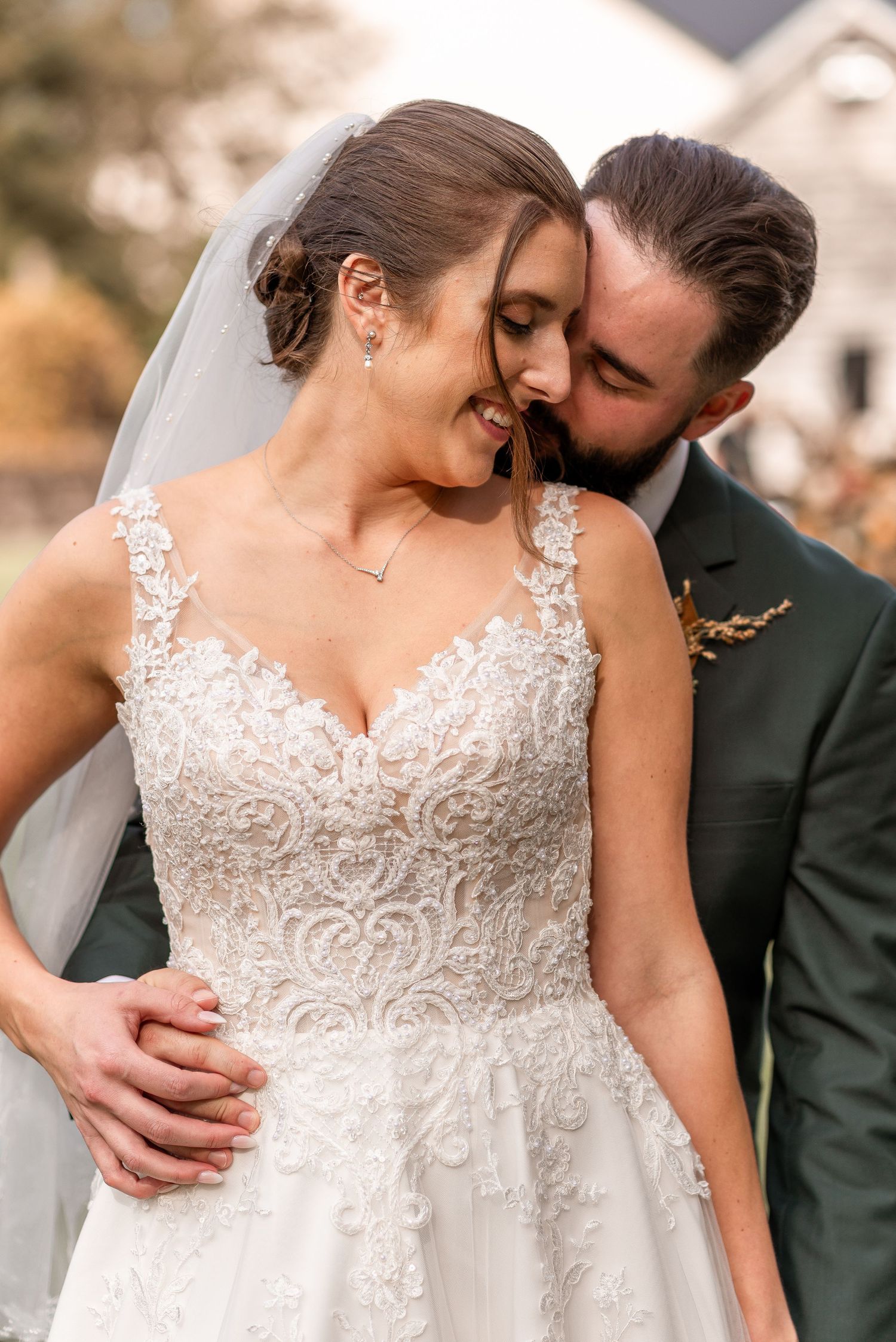 Groom in dark green suit nuzzling smiling bride in lace wedding gown outdoors at fall wedding.