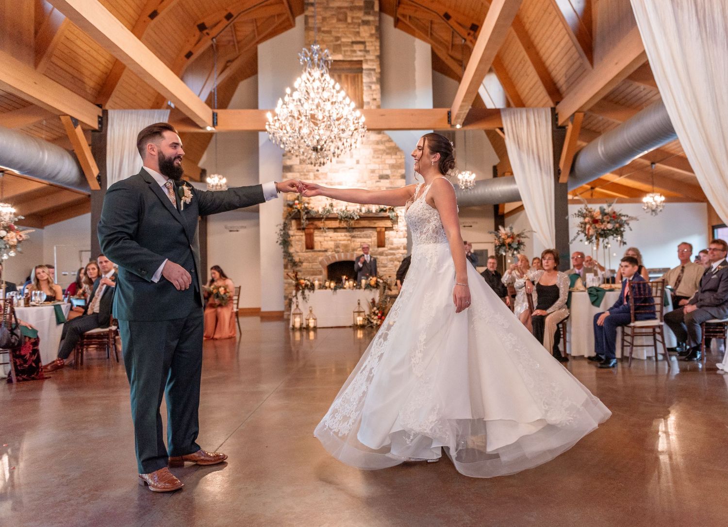 Bride and groom sharing first dance in rustic wooden venue with chandelier and stone fireplace backdrop.