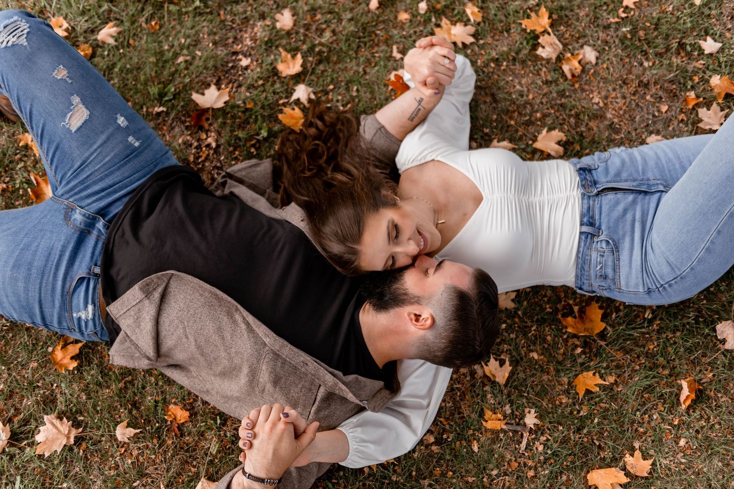 Couple lying on grass surrounded by autumn leaves, sharing a tender moment during engagement photos.