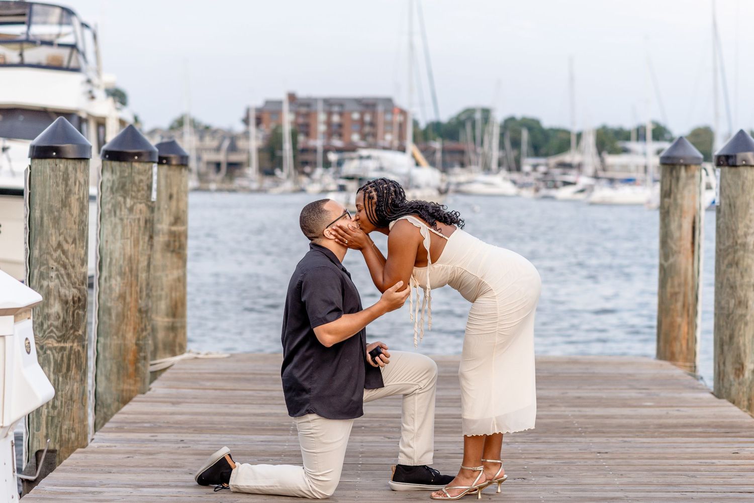 Couple sharing a kiss on a marina dock after a marriage proposal with boats in the background.