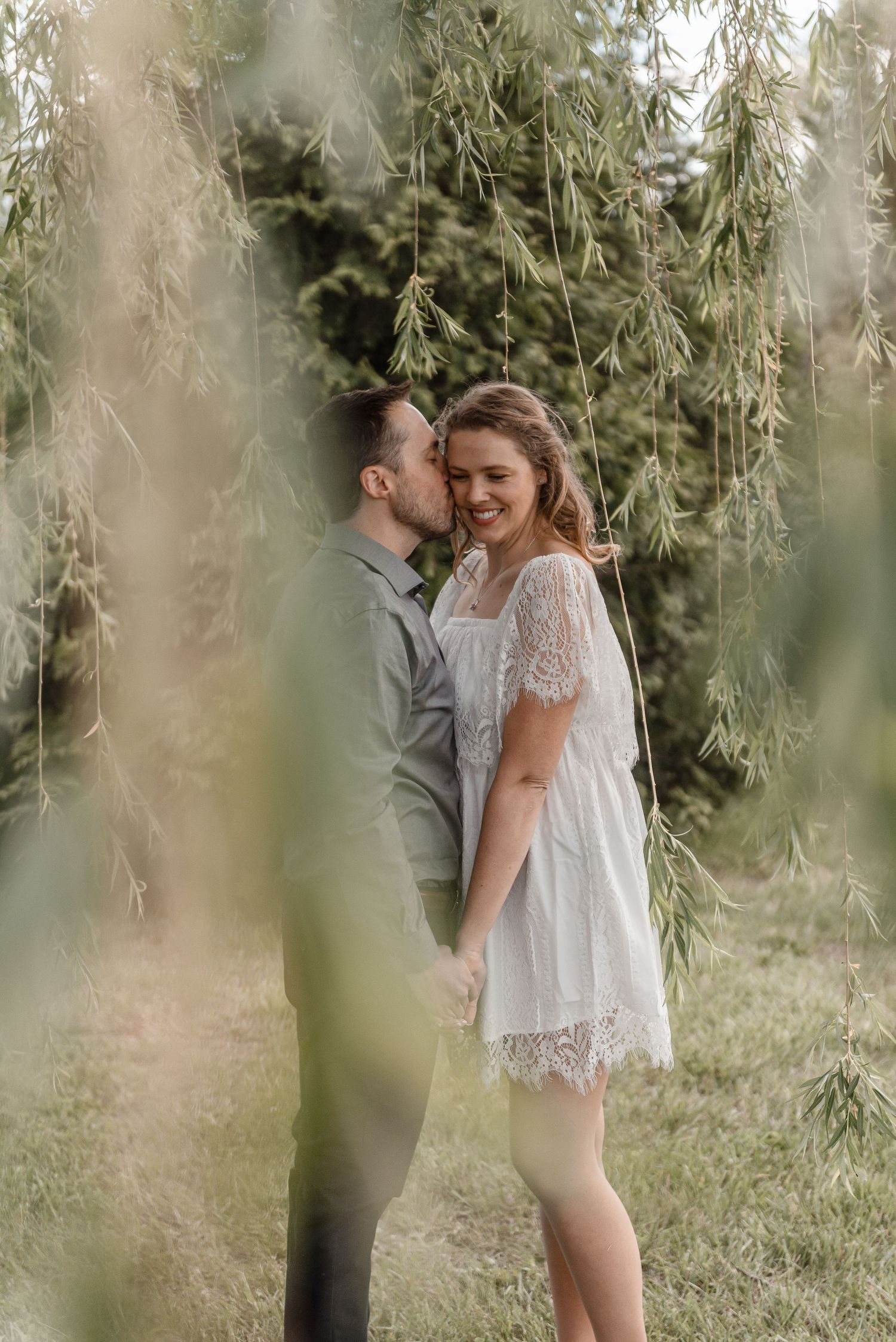 Man kissing woman on cheek as she smiles under hanging willow tree branches outdoors.