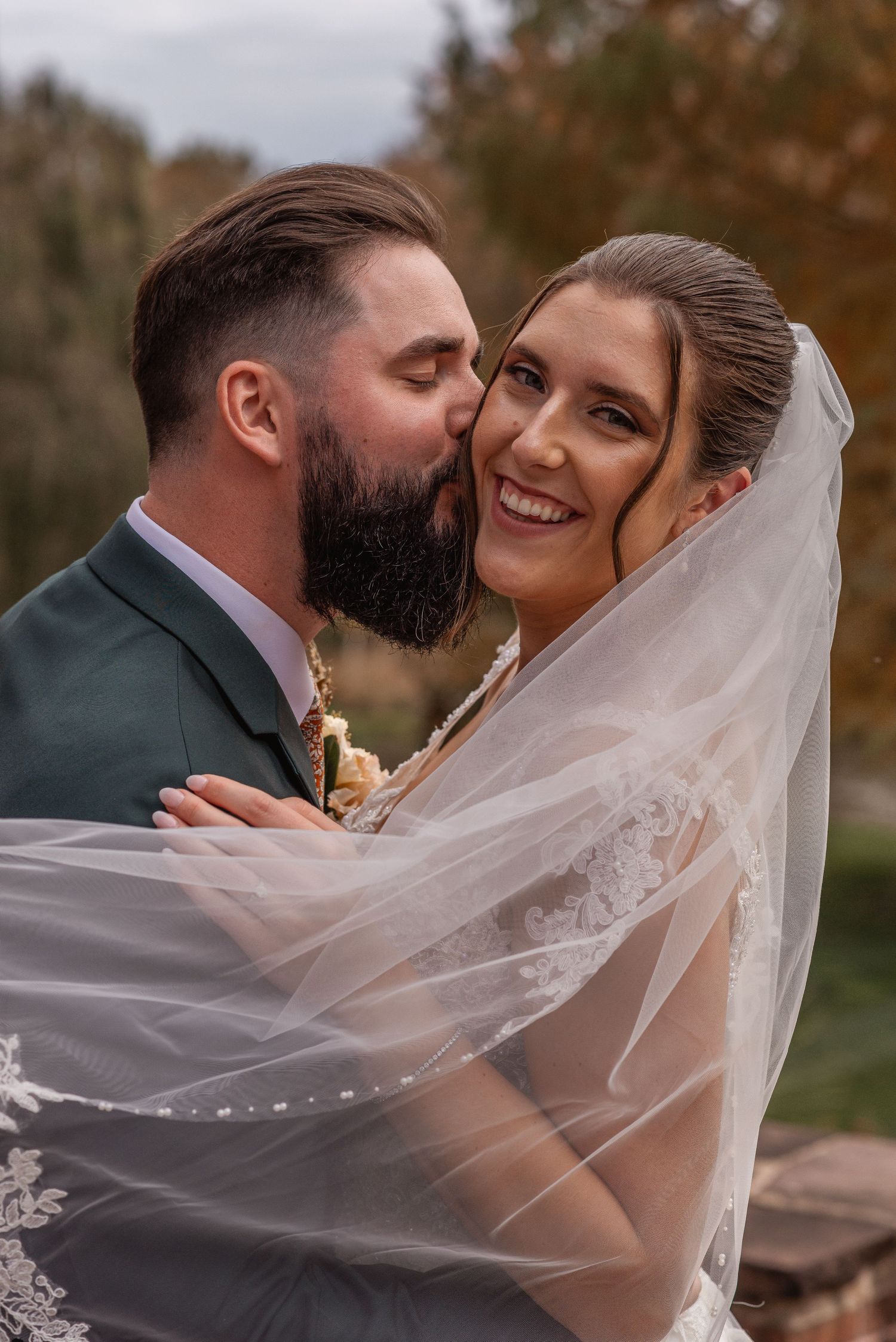 Bearded groom in green suit kissing smiling bride on cheek, her veil flowing around them in autumn setting.