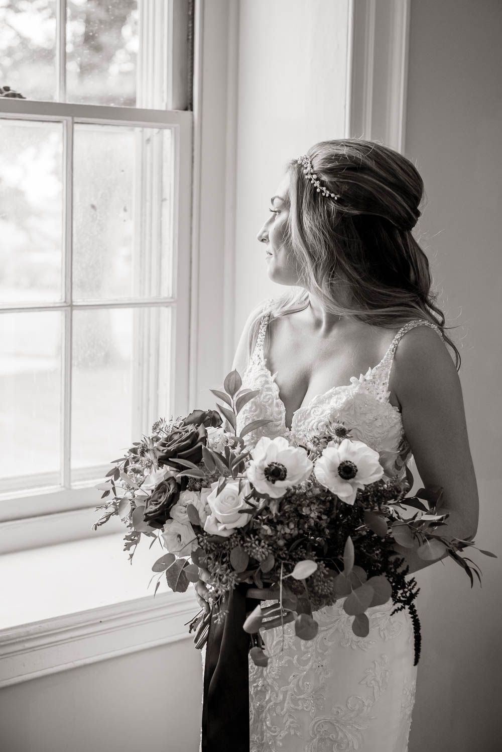 Elegant bride in lace gown holds floral bouquet with anemones while gazing out window in black and white.