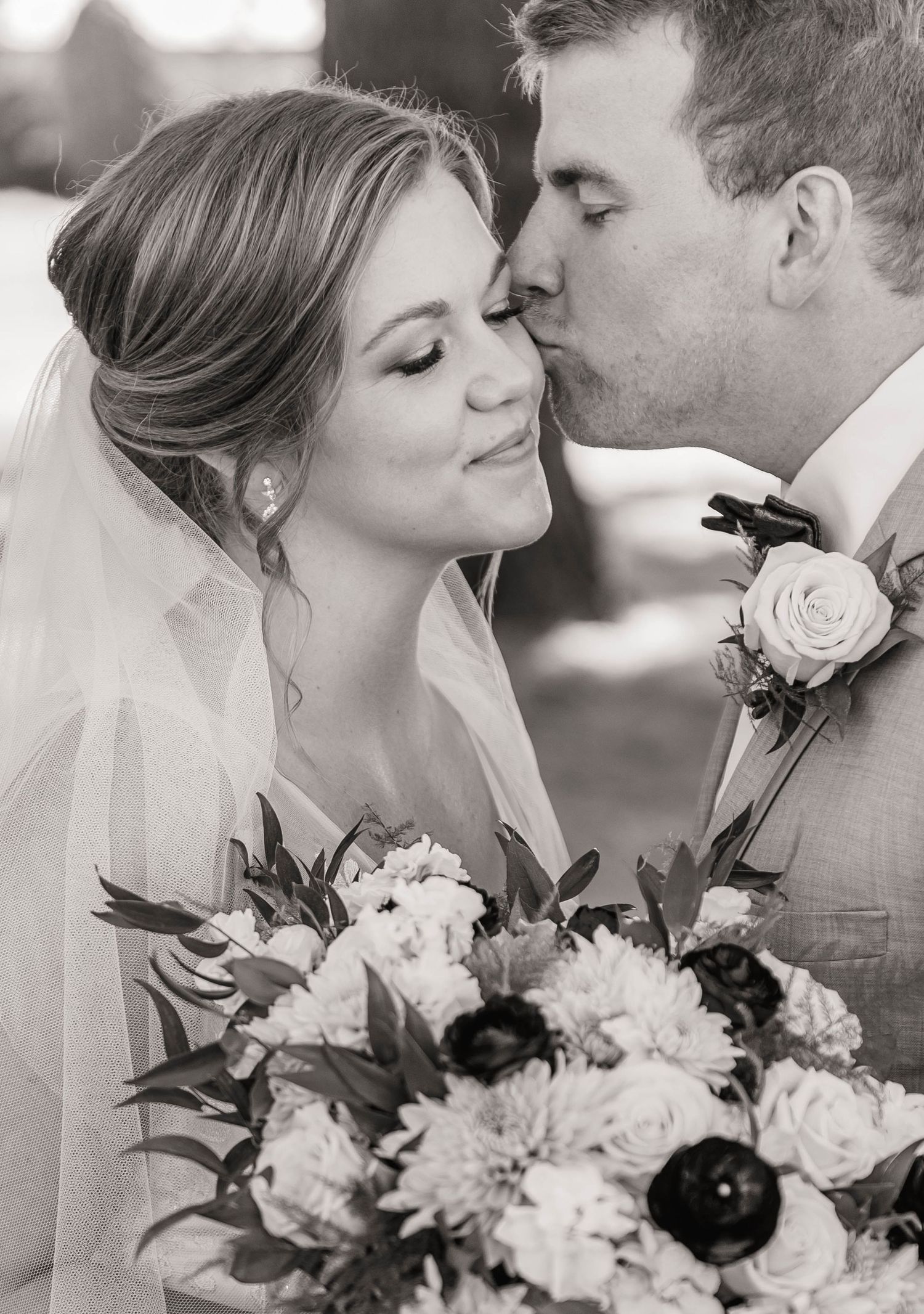 Black and white photo of groom kissing bride on cheek as she smiles holding a lush bouquet.