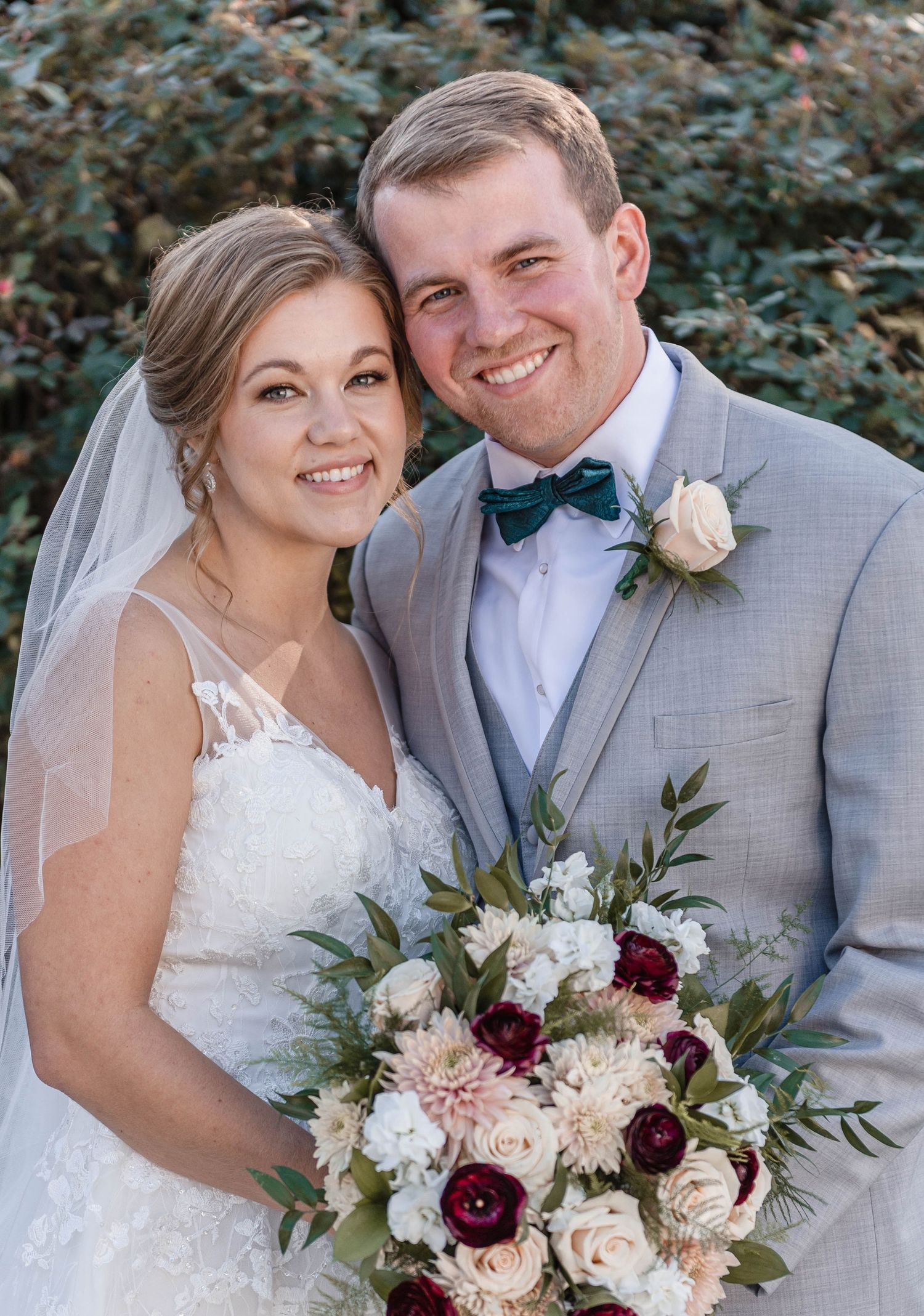 Happy bride and groom smiling together outdoors, holding burgundy and blush bouquet.