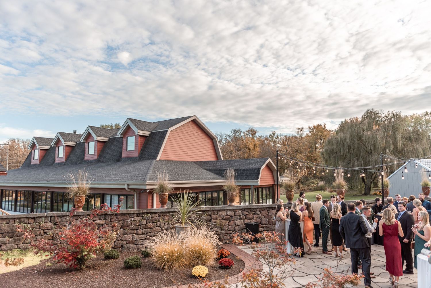 Guests mingling outside a charming stone and red barn wedding venue under dramatic cloudy autumn sky.