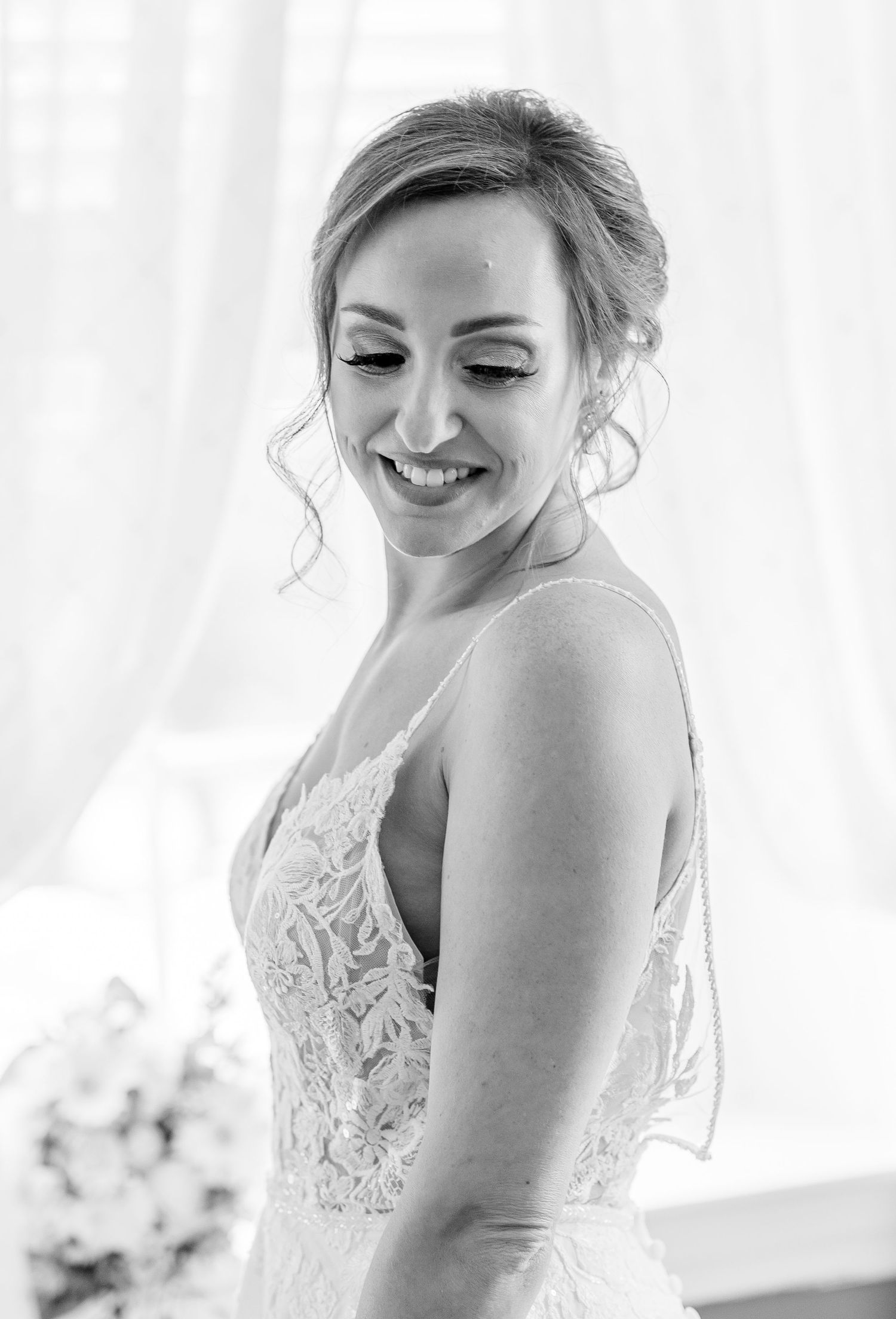 Black and white portrait of a glowing bride in a lace gown smiling softly with eyes downcast near a bright window.