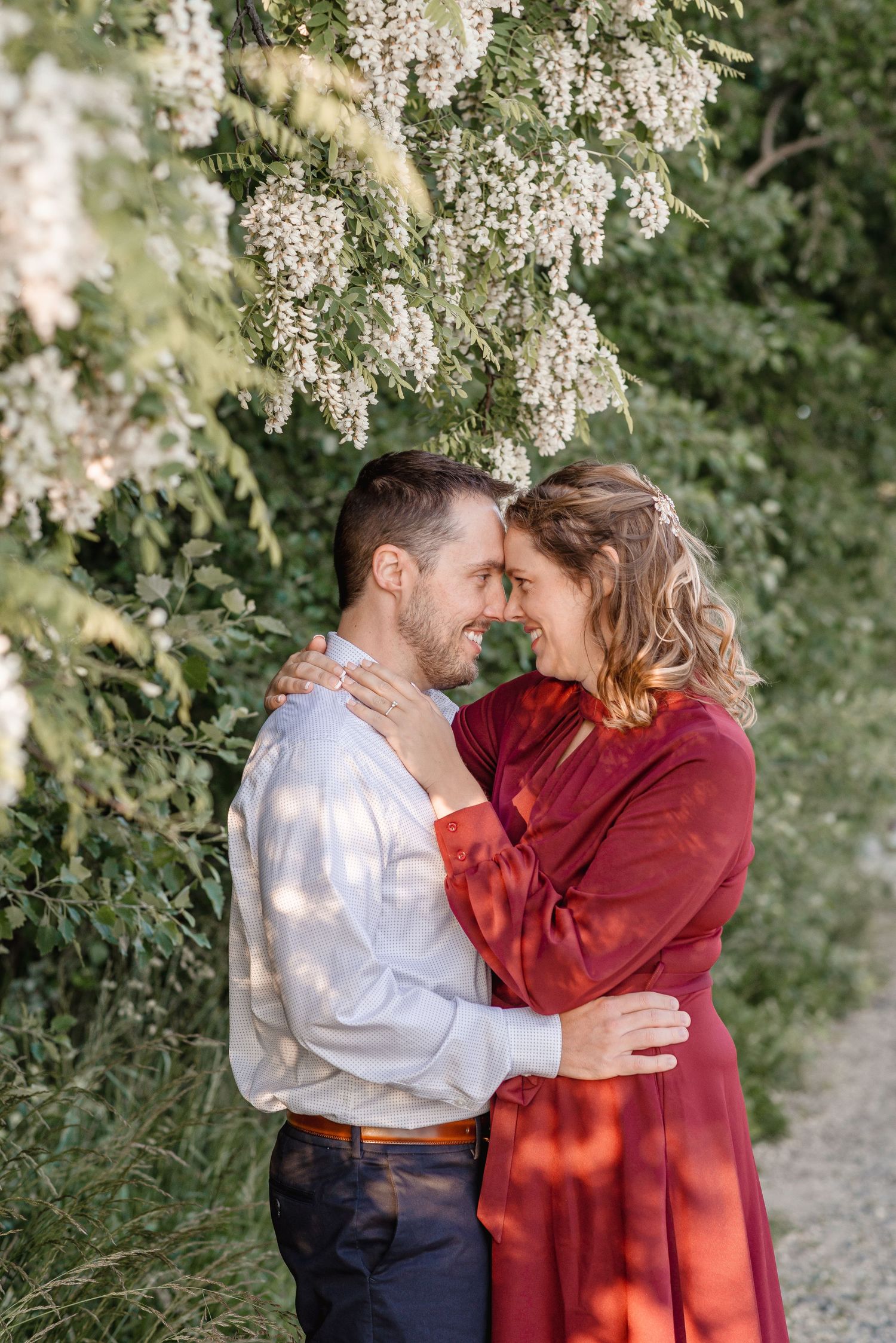 Couple embracing and touching foreheads under blooming white flowers in a lush garden setting.