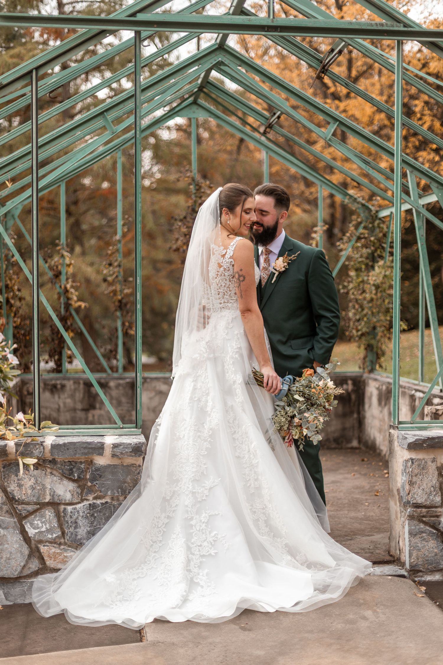 A bride and groom share a romantic moment in front of a green iron greenhouse in autumn surroundings.
