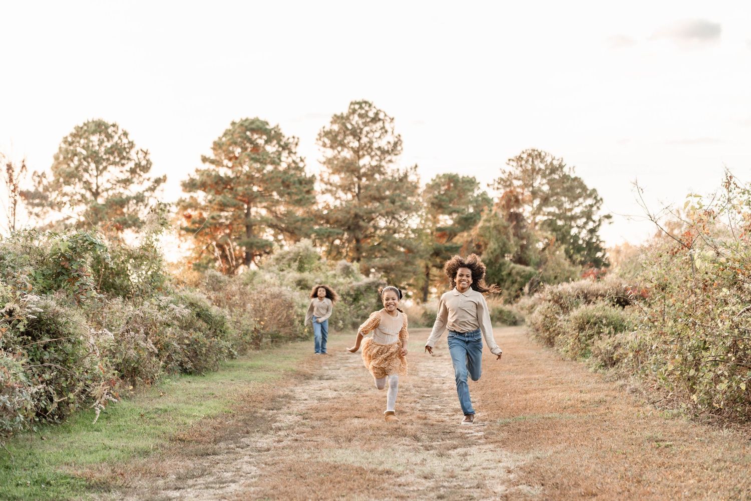 Three children running joyfully down a sunlit dirt path surrounded by golden autumn foliage and tall pine trees.