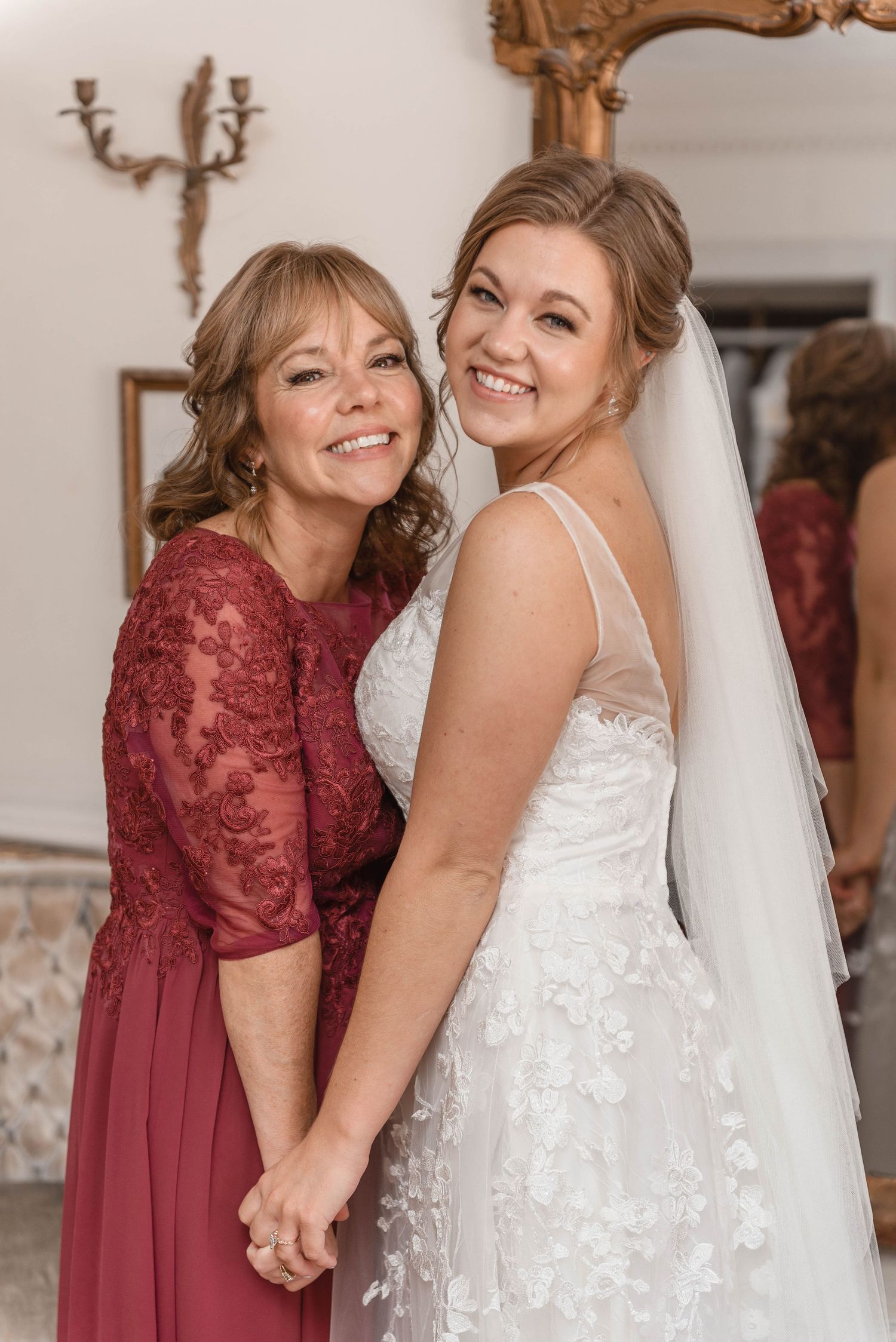 Smiling bride in white lace gown poses with mother in burgundy dress on wedding day.