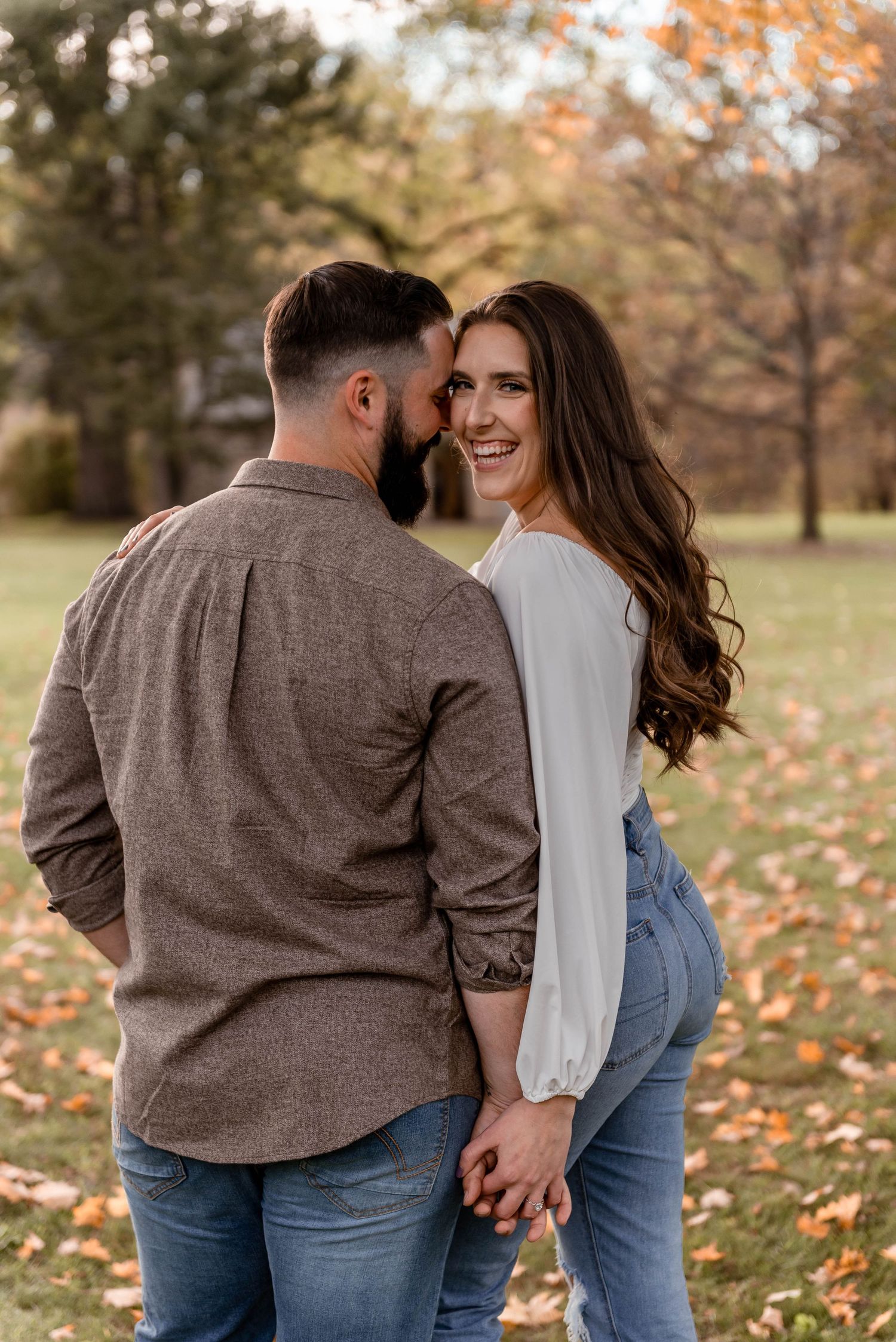 Couple holding hands in an autumn park, woman smiling over her shoulder during fall engagement photo session.