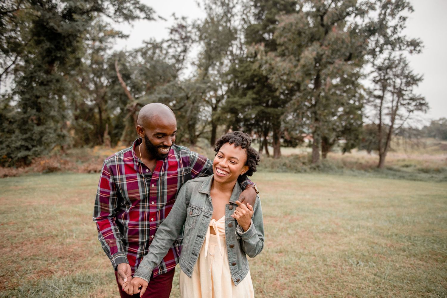 A joyful couple walks hand in hand through a green field during an outdoor engagement photo session.