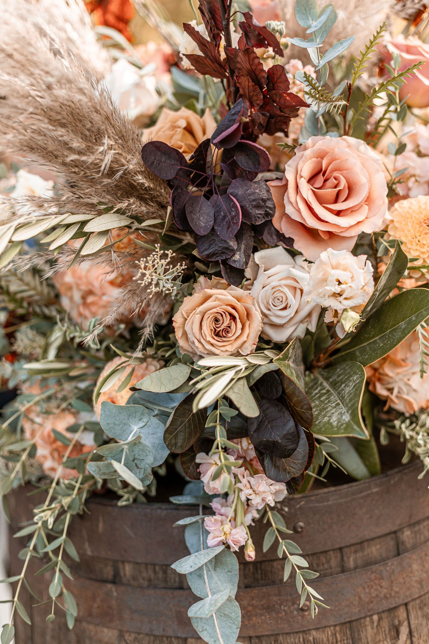 Elegant wedding floral arrangement featuring peach roses, pampas grass, eucalyptus, and burgundy blooms on a wooden barrel.