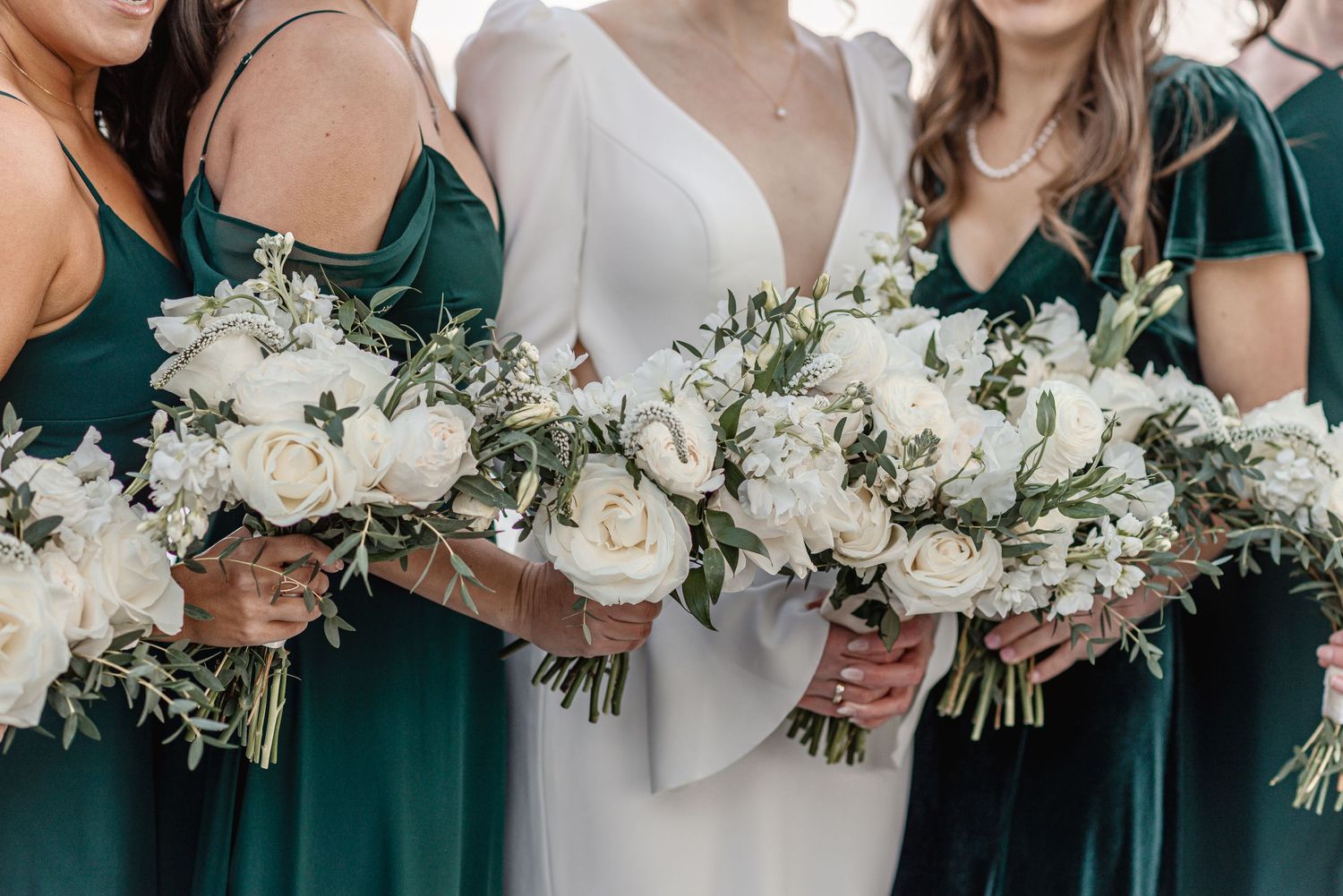 Bride flanked by bridesmaids in emerald green dresses holding white rose and greenery bouquets.