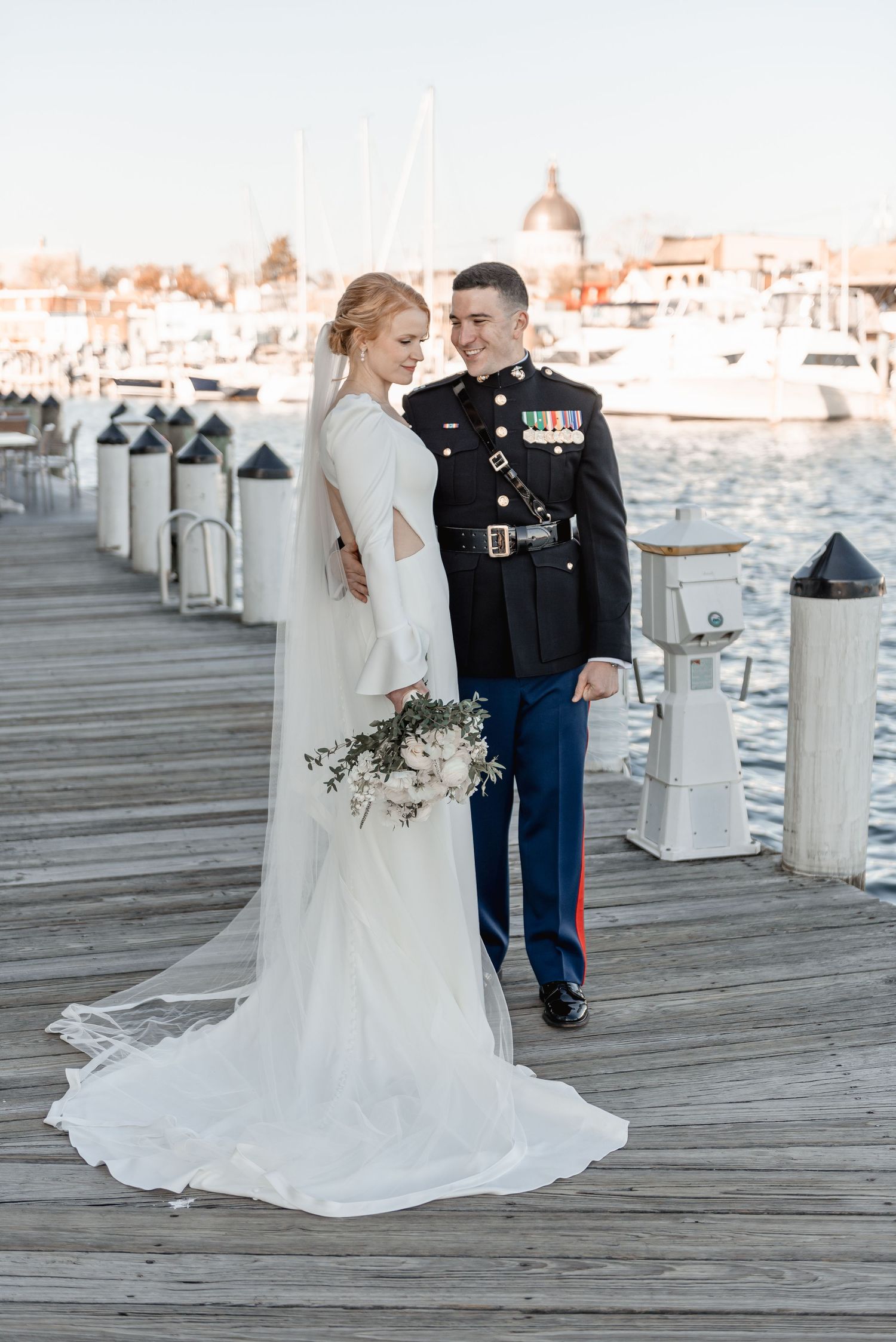 Military groom in dress uniform and bride holding bouquet walk hand-in-hand on a marina dock at sunset.