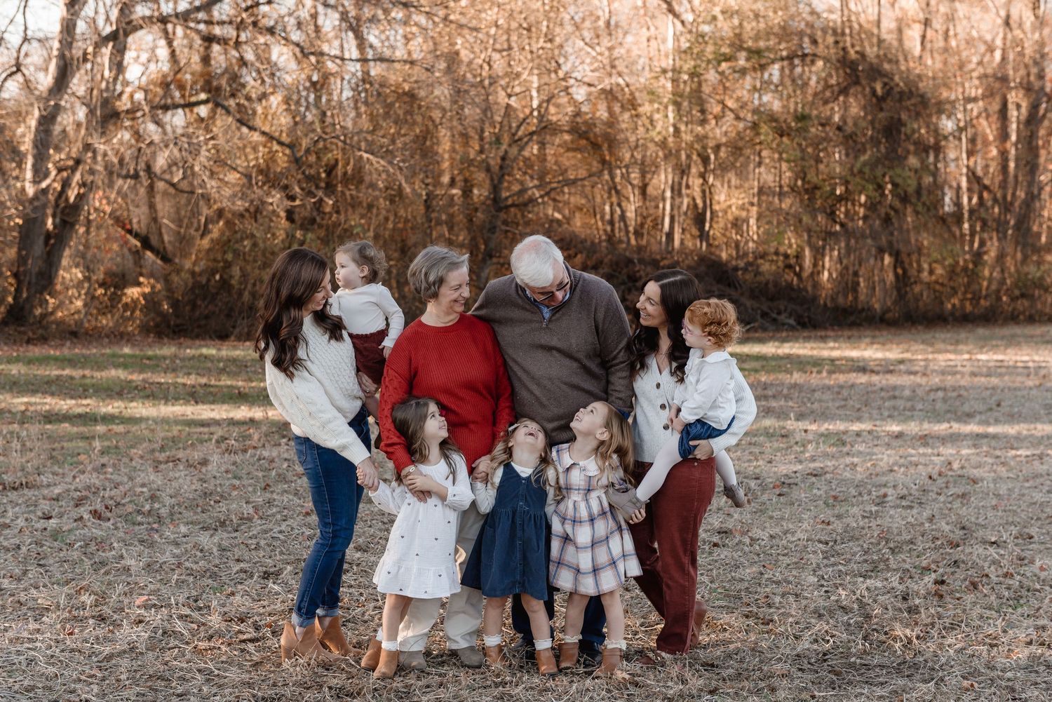 Multi-generational family group hugging outdoors in golden autumn light among bare trees.