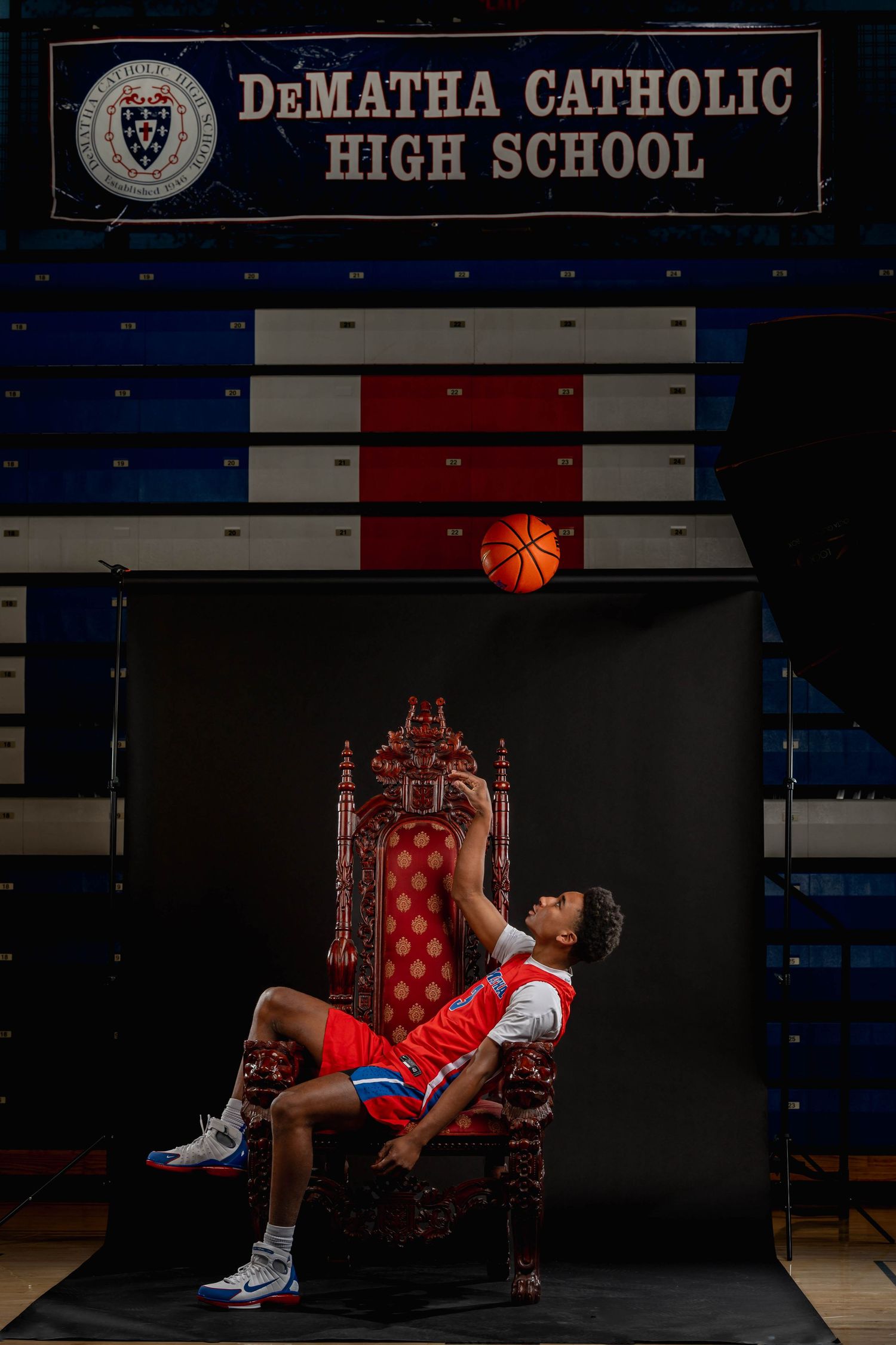 A young basketball player sits on a throne at DeMatha Catholic High School, tossing a basketball in the air.