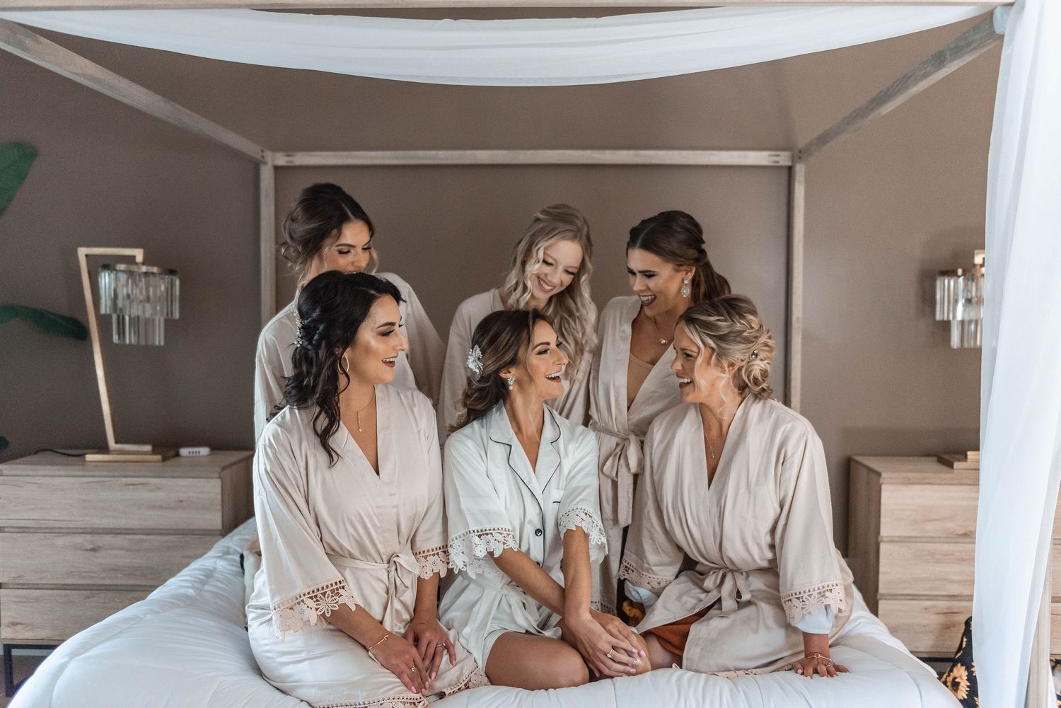 Bride and bridesmaids laughing together in matching robes on a bed during wedding morning preparations.