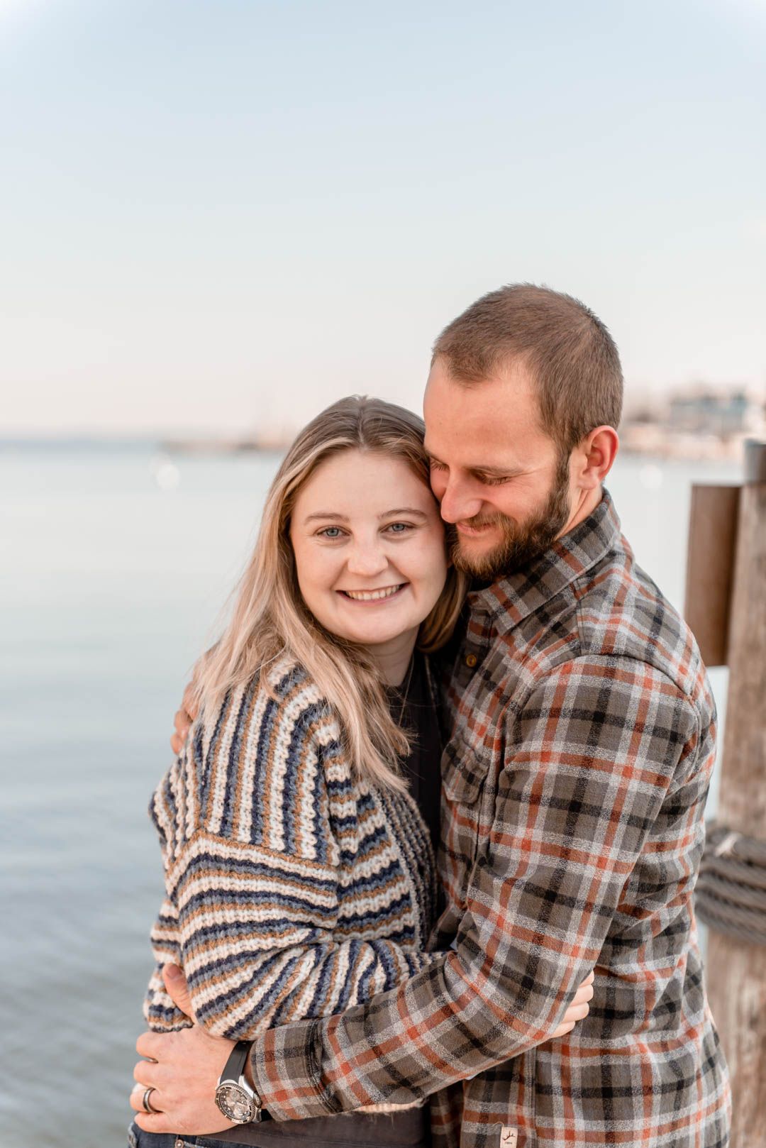 Smiling couple embracing near a waterfront dock, woman in striped sweater and man in plaid shirt.